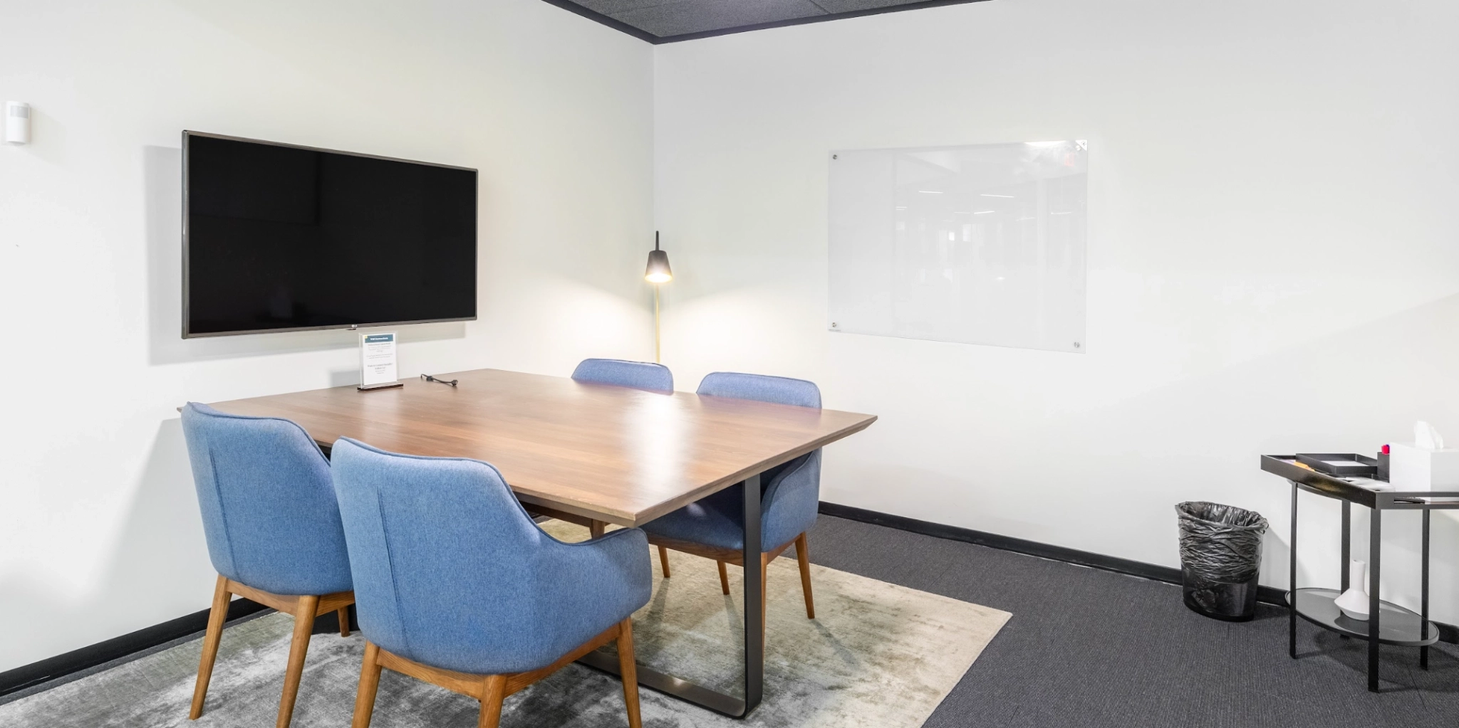 A small meeting room with a wooden table, four blue chairs, a wall-mounted TV, a whiteboard, a floor lamp, and a wastebasket. This well-equipped workspace also includes a tissue box and sanitizer on a side table.