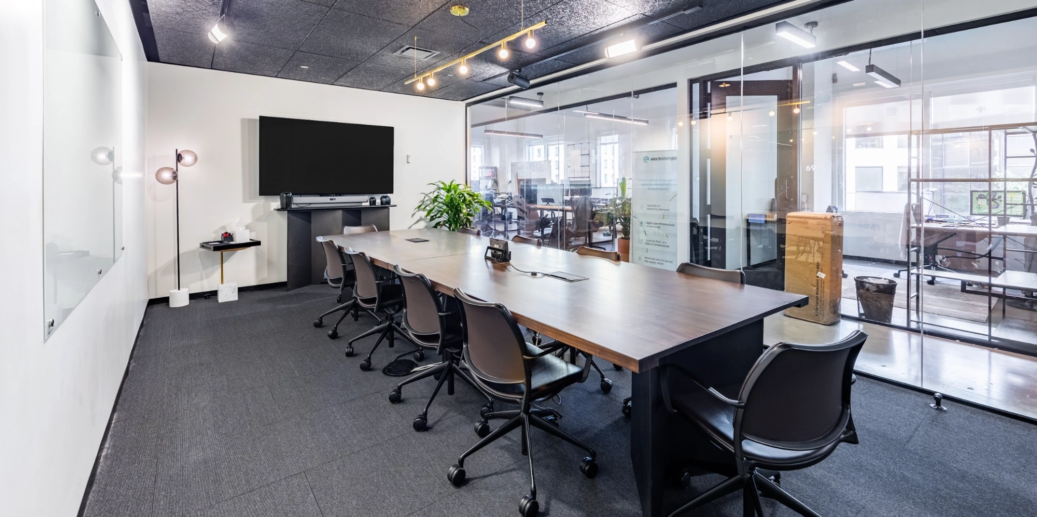 Une salle de conférence moderne avec une longue table, plusieurs chaises roulantes, un grand écran au mur et des cloisons en verre la séparant de l'espace bureau de coworking.