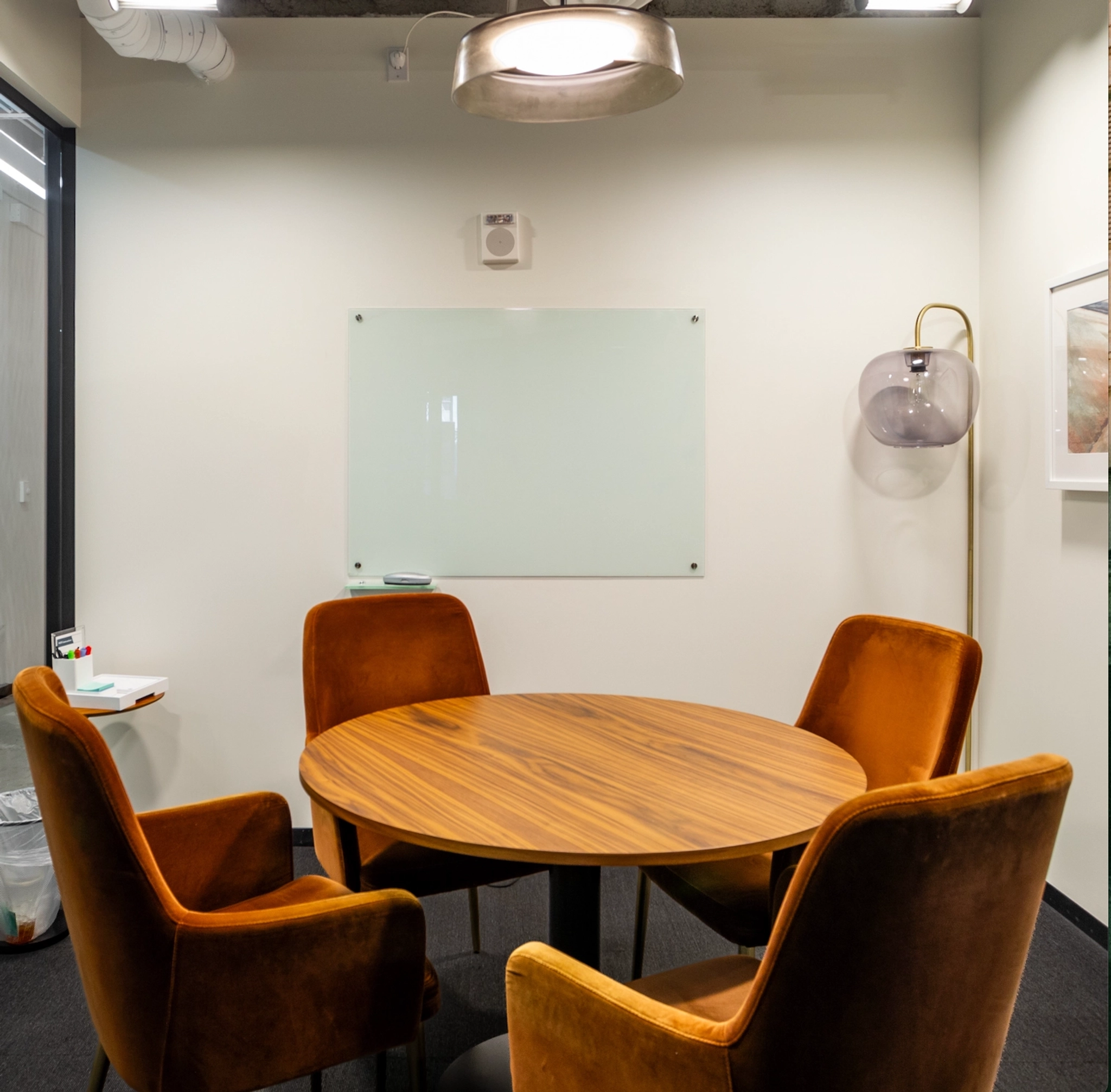 Een warme werkruimte met een ronde houten tafel omringd door vier oranje gestoffeerde stoelen. Deze intieme vergaderruimte wordt geaccentueerd met een aan de muur gemonteerd whiteboard en zacht verlicht door een enkele wandlamp in de hoek.