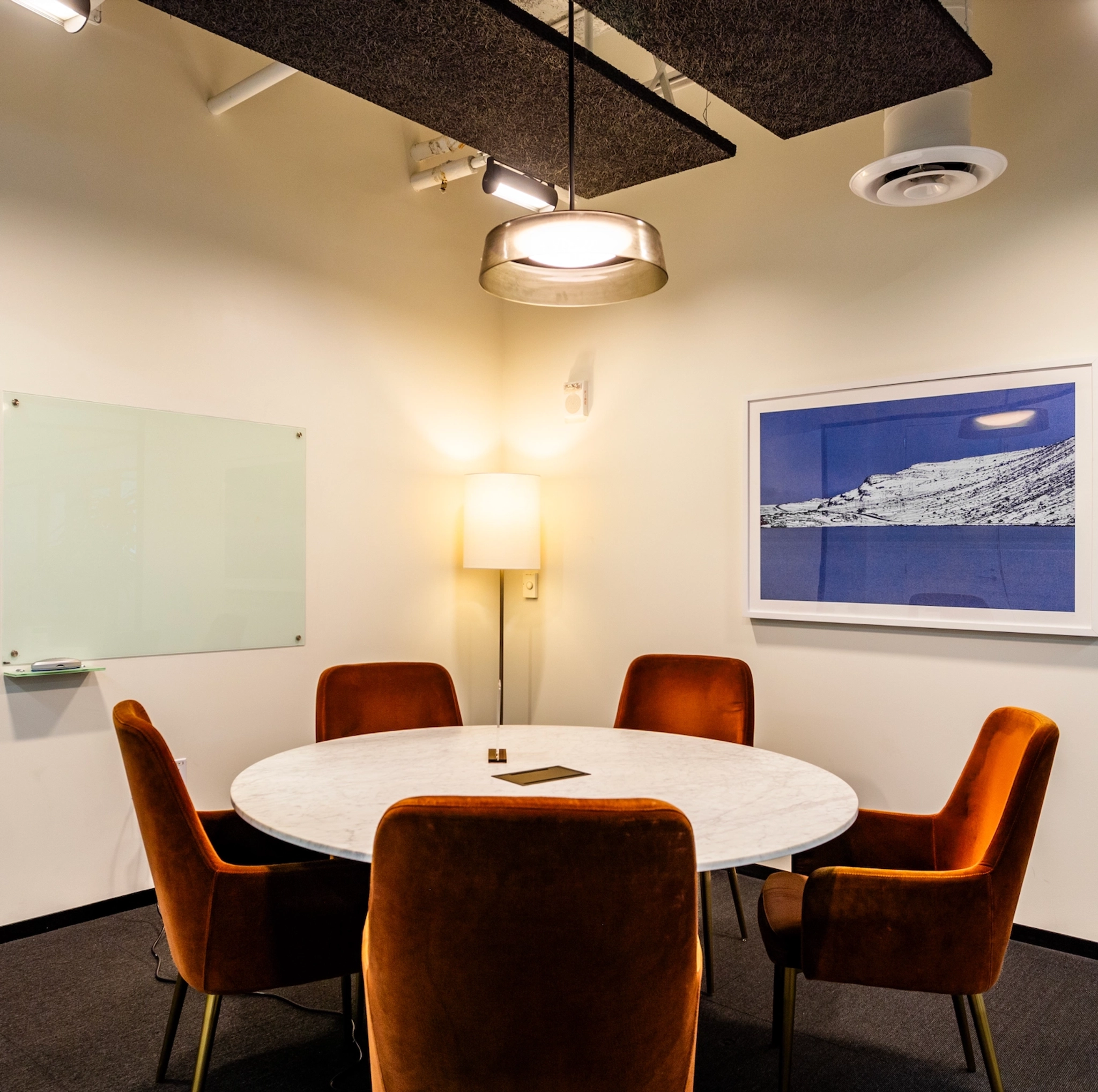 A small coworking meeting room in San Francisco features a round table surrounded by four orange chairs. There's a whiteboard, a floor lamp, and a framed picture on the wall. A ceiling light hangs above the table, creating an ideal space for collaboration.