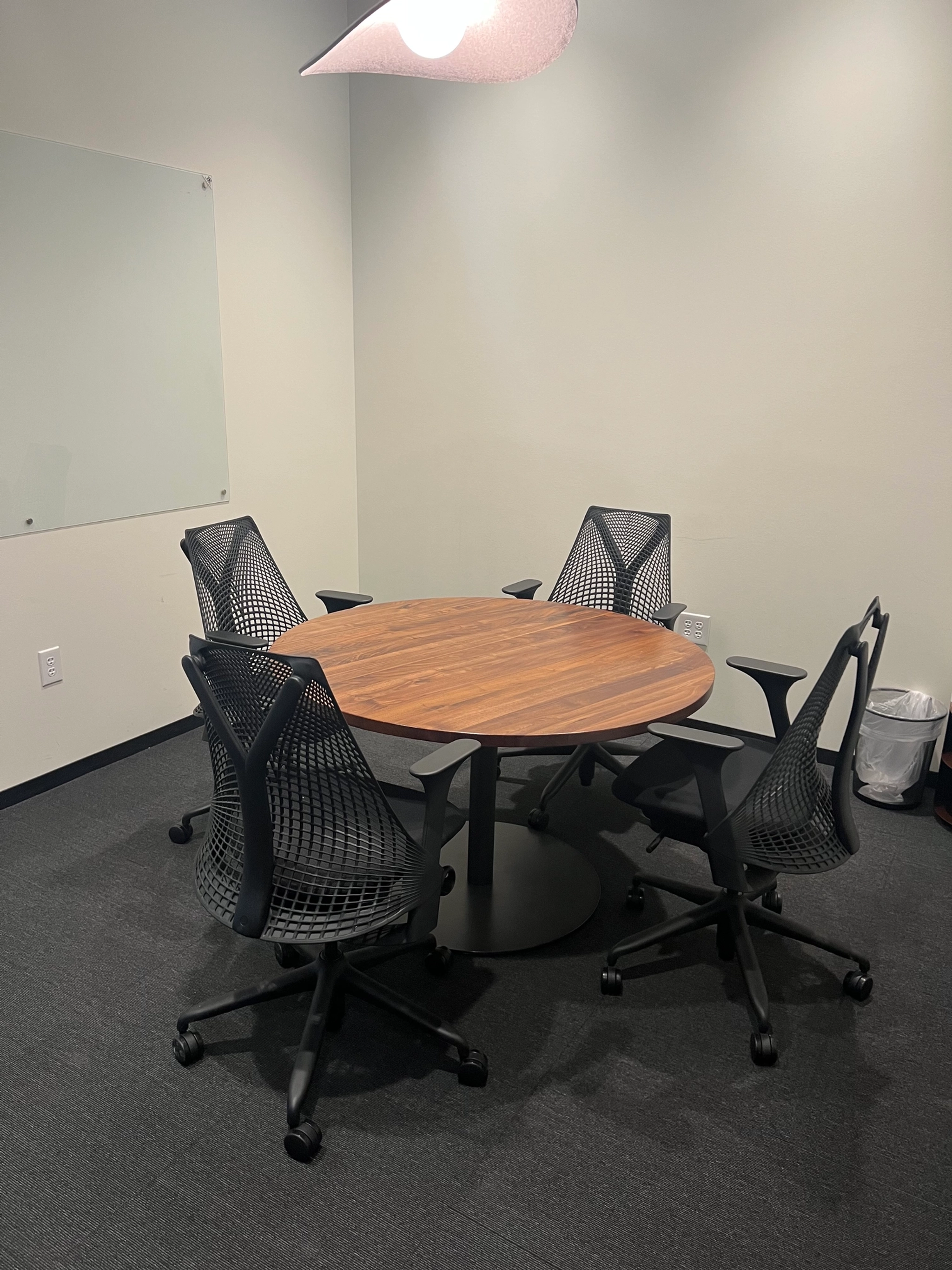 A small meeting room in a Dallas coworking space features a round wooden table surrounded by four black mesh office chairs, with a whiteboard on the wall and a trash can in the corner.