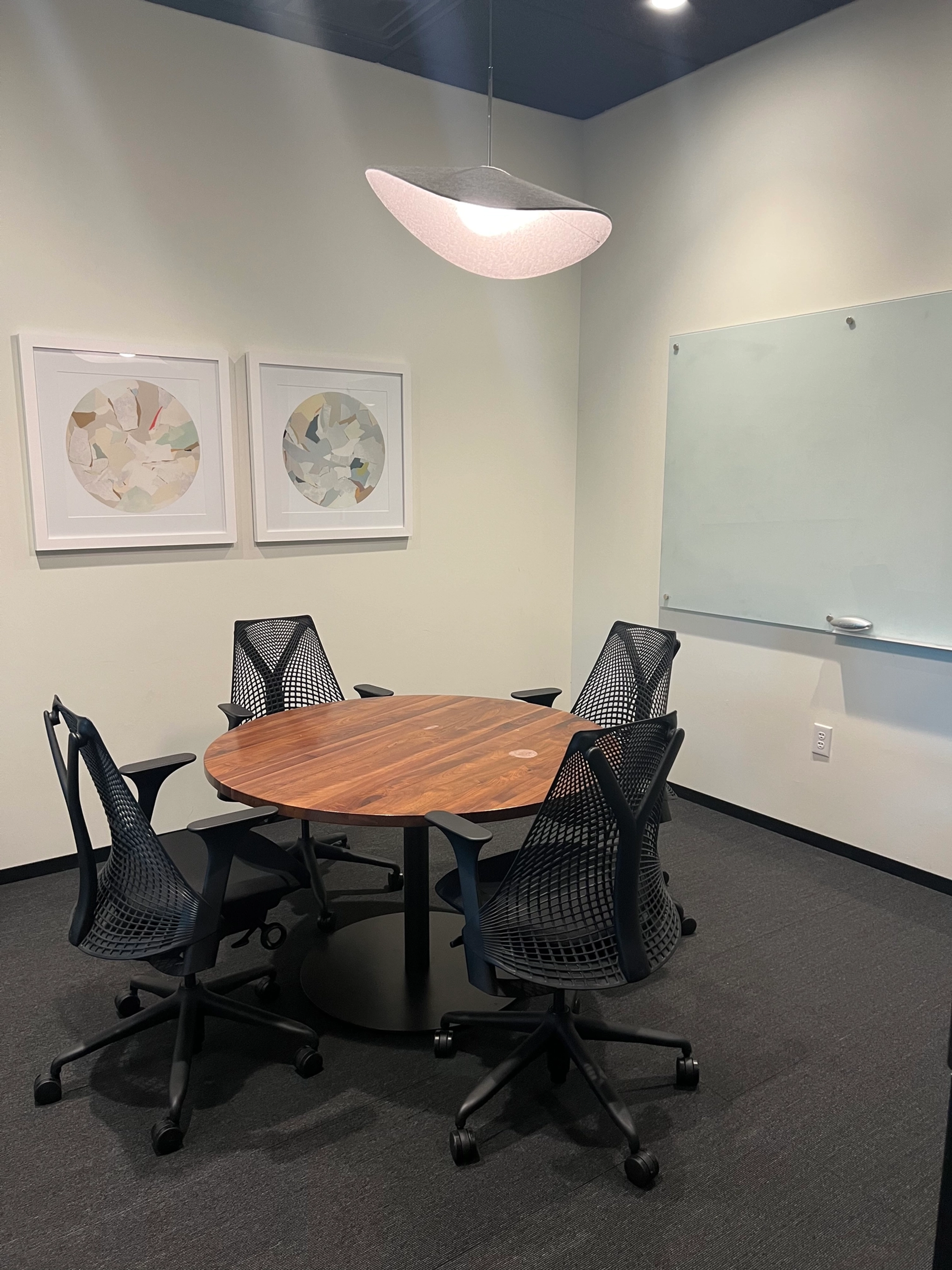 A small conference room in Dallas featuring a round wooden table, four black mesh office chairs, a whiteboard, and two framed abstract artworks on the wall. Perfect for coworking sessions or meetings in a dynamic workspace setting.