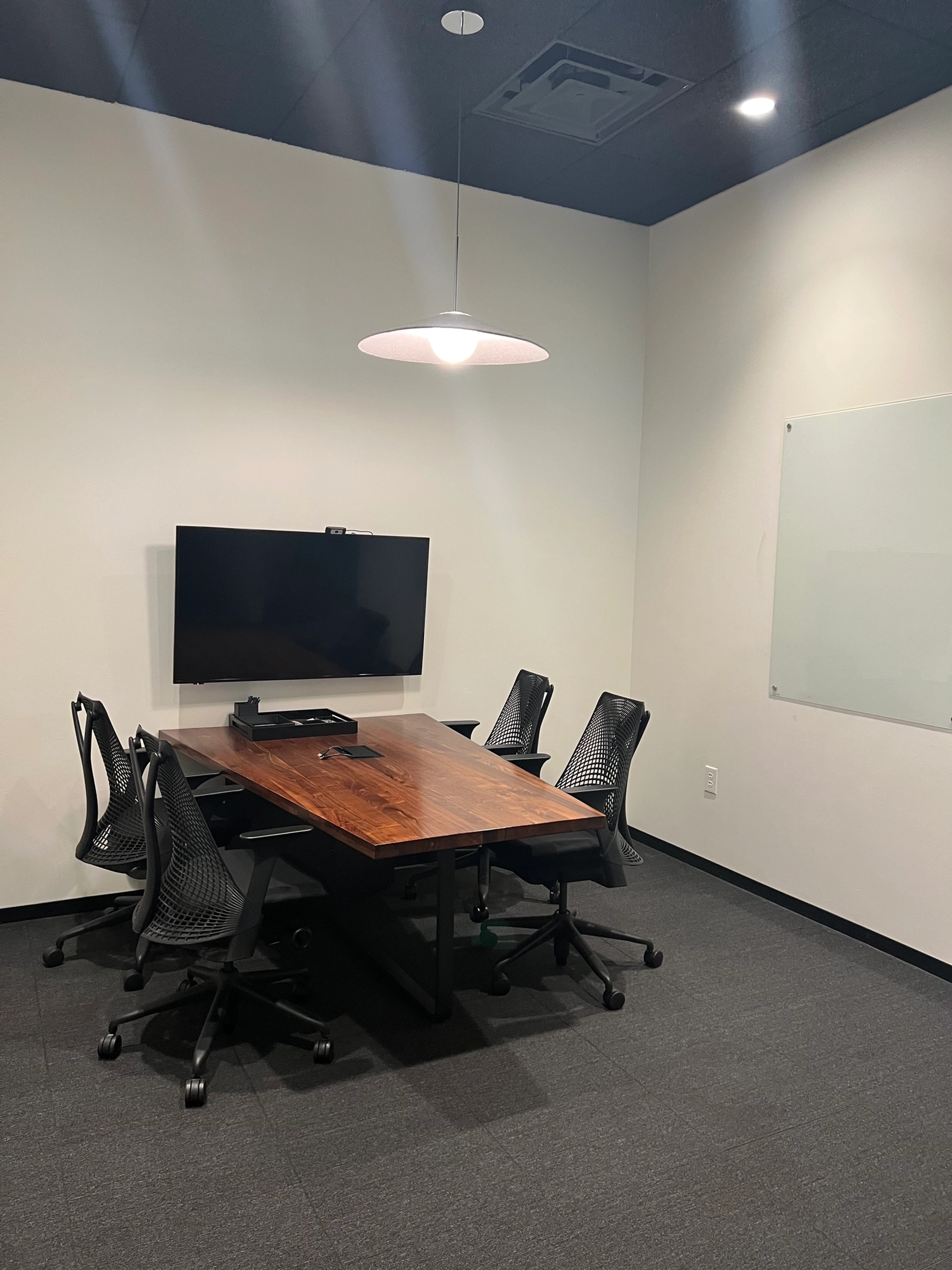 A small coworking meeting room with a wooden table, four office chairs, a wall-mounted monitor, a whiteboard, and a ceiling light. The walls and carpet are neutral in color.