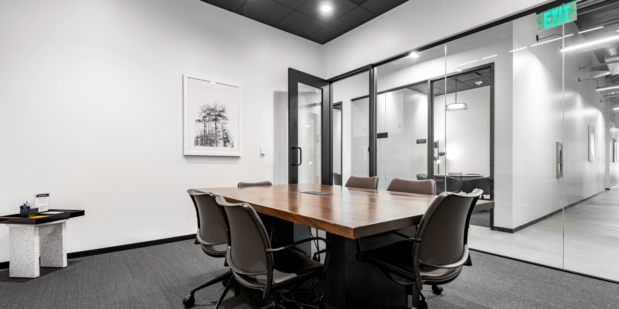 Modern meeting room in Walnut Creek with a wooden table, four chairs, and glass walls. A framed black-and-white photo hangs on a white wall, adding a touch of elegance. An exit sign is visible above the door, ensuring safety in the coworking space.