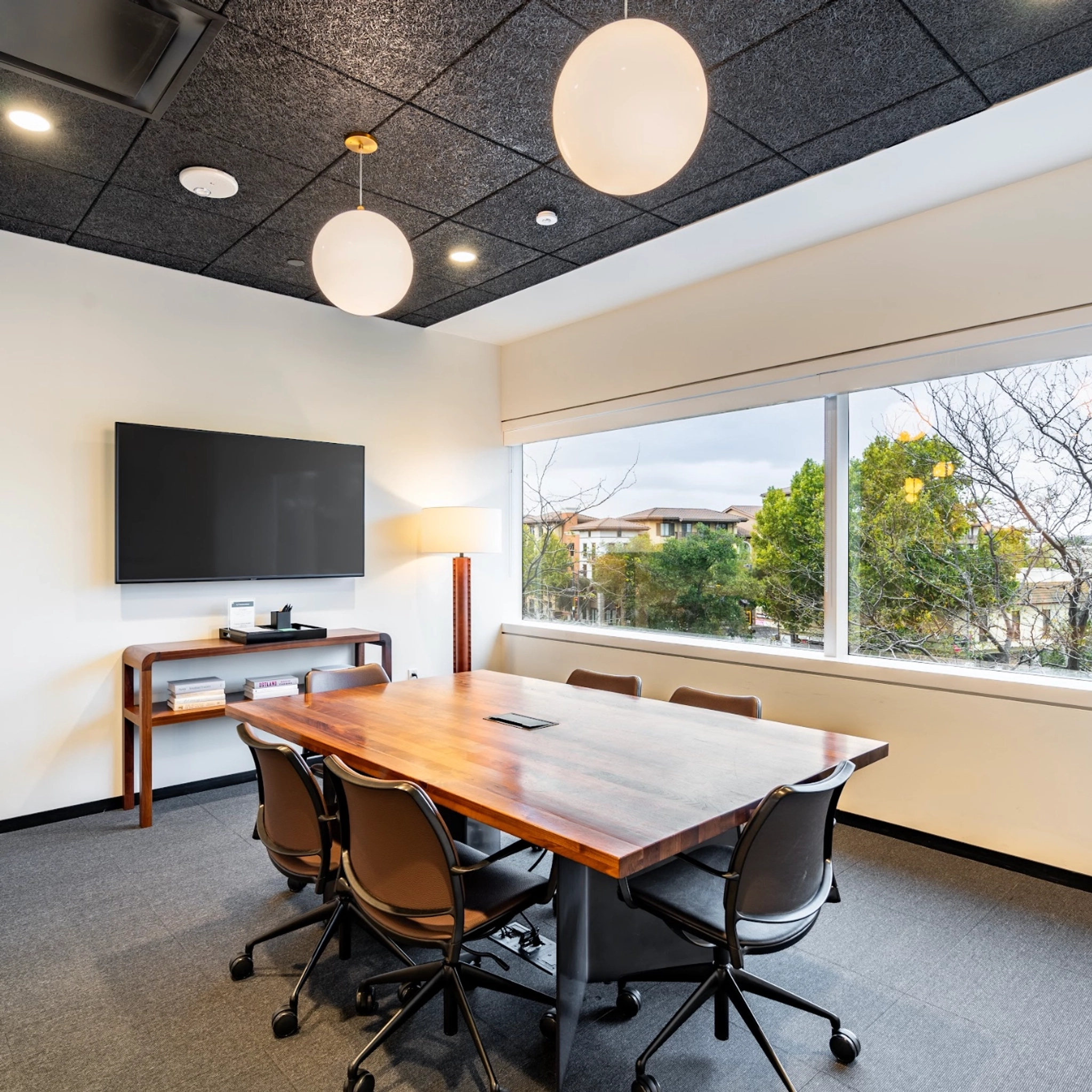 A modern coworking space features a conference room with a wooden table, six chairs, a wall-mounted TV, and a wooden sideboard. Large windows overlook the trees outside, while three round ceiling lights illuminate the perfect setting for team meetings.