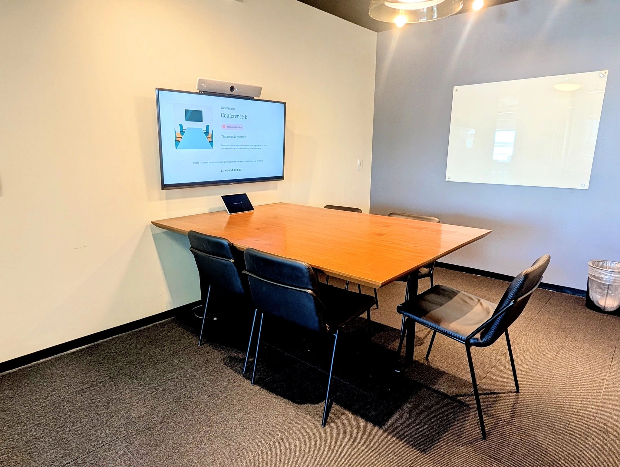 Una pequeña sala de reuniones de coworking en Washington con una mesa de madera, seis sillas, una pantalla en la pared que muestra una presentación, una pizarra blanca y una papelera.