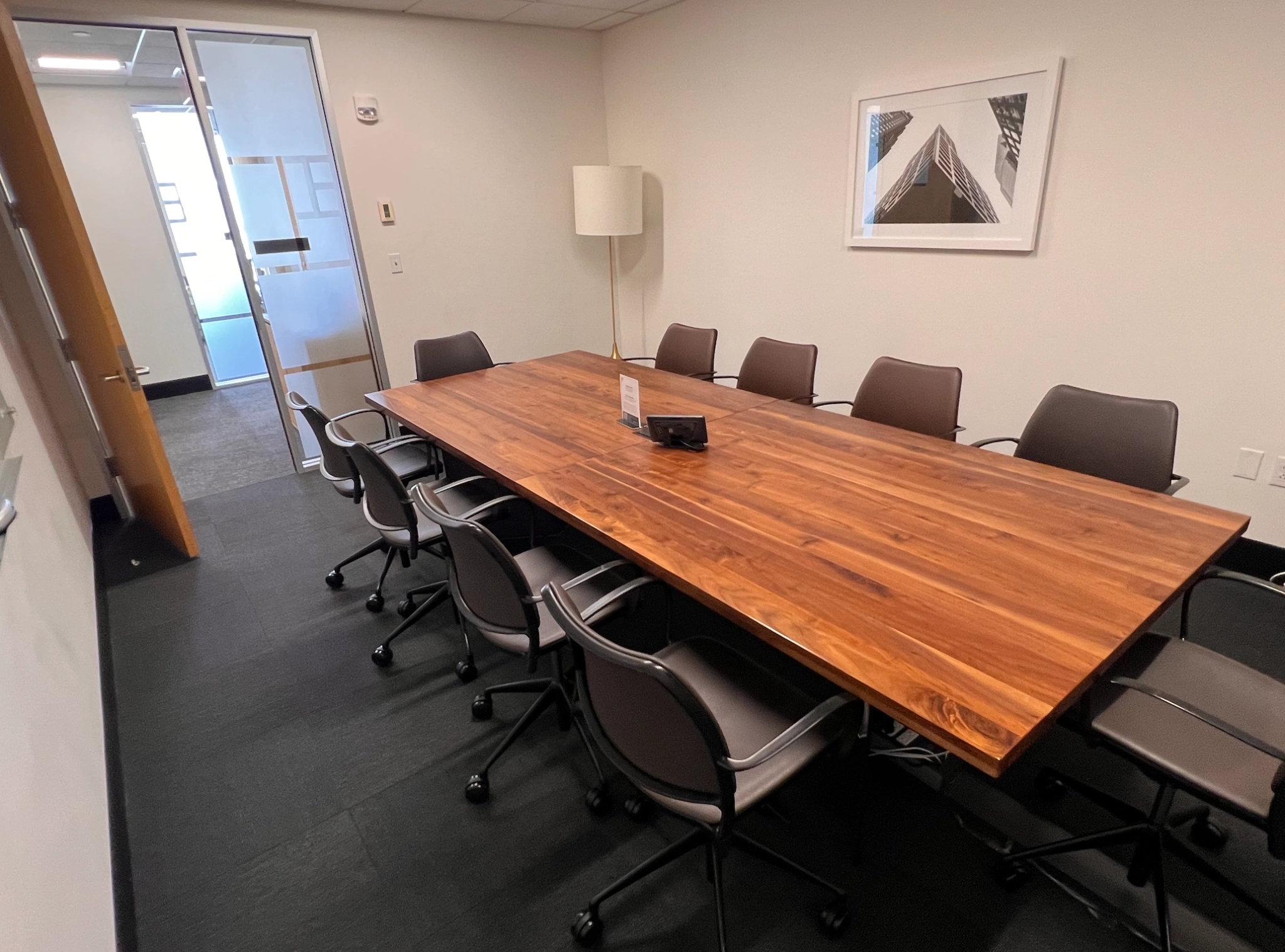 Une salle de réunion à Boston avec une table en bois, dix chaises, une lampe, un tableau encadré au mur et une porte vitrée ouvrant sur l'espace de travail adjacent.
