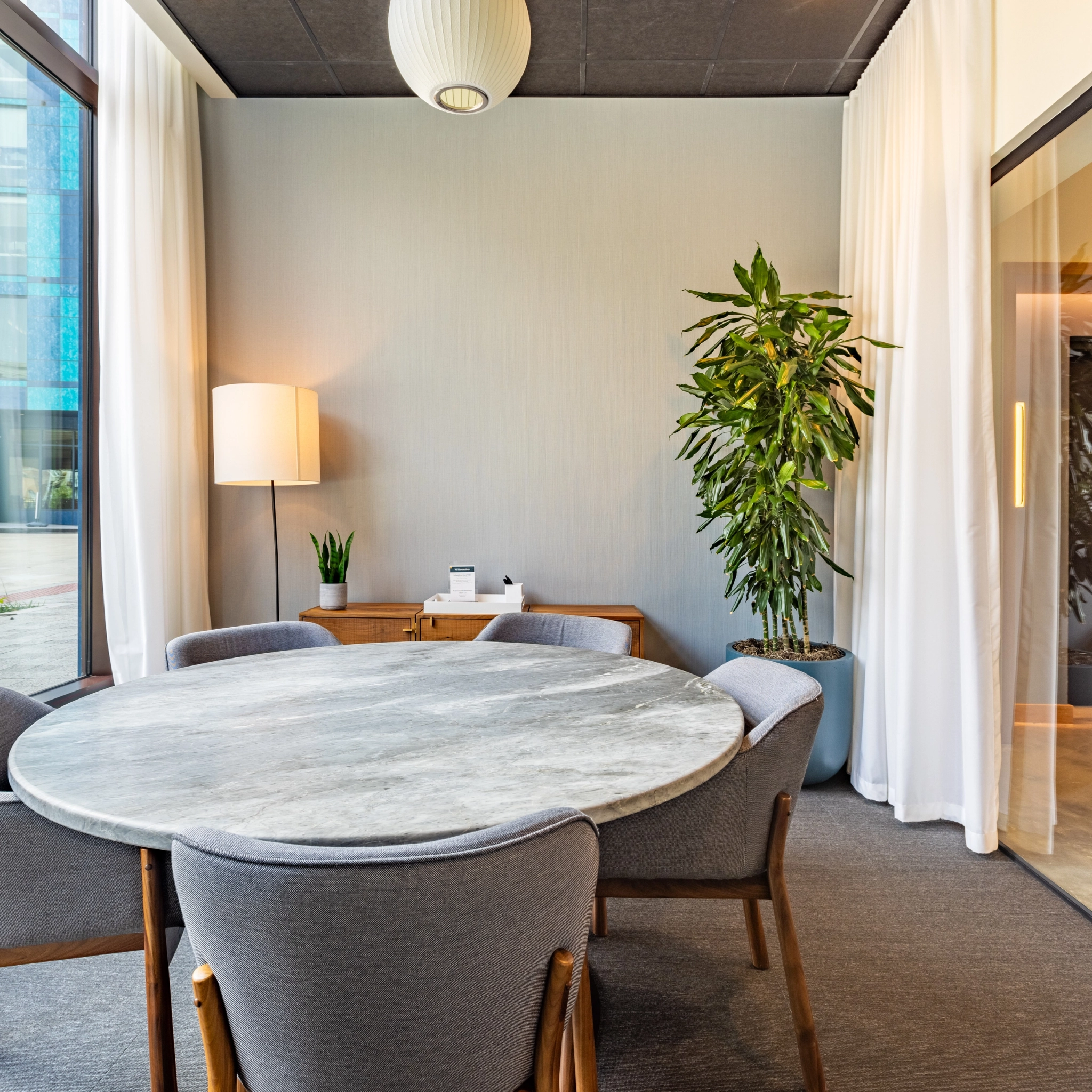 A modern meeting room in a Washington coworking space features a round marble table, four chairs, potted plants, a floor lamp, and a side cabinet.