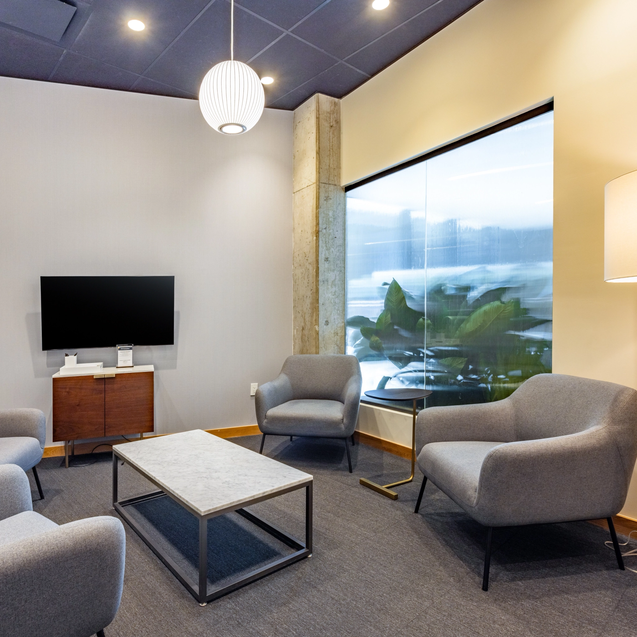 A modern coworking waiting area in Washington features grey armchairs, a rectangular coffee table, a flat-screen TV on a wooden stand, and a large window with outdoor views. A pendant light hangs from the ceiling, illuminating the stylish workspace.