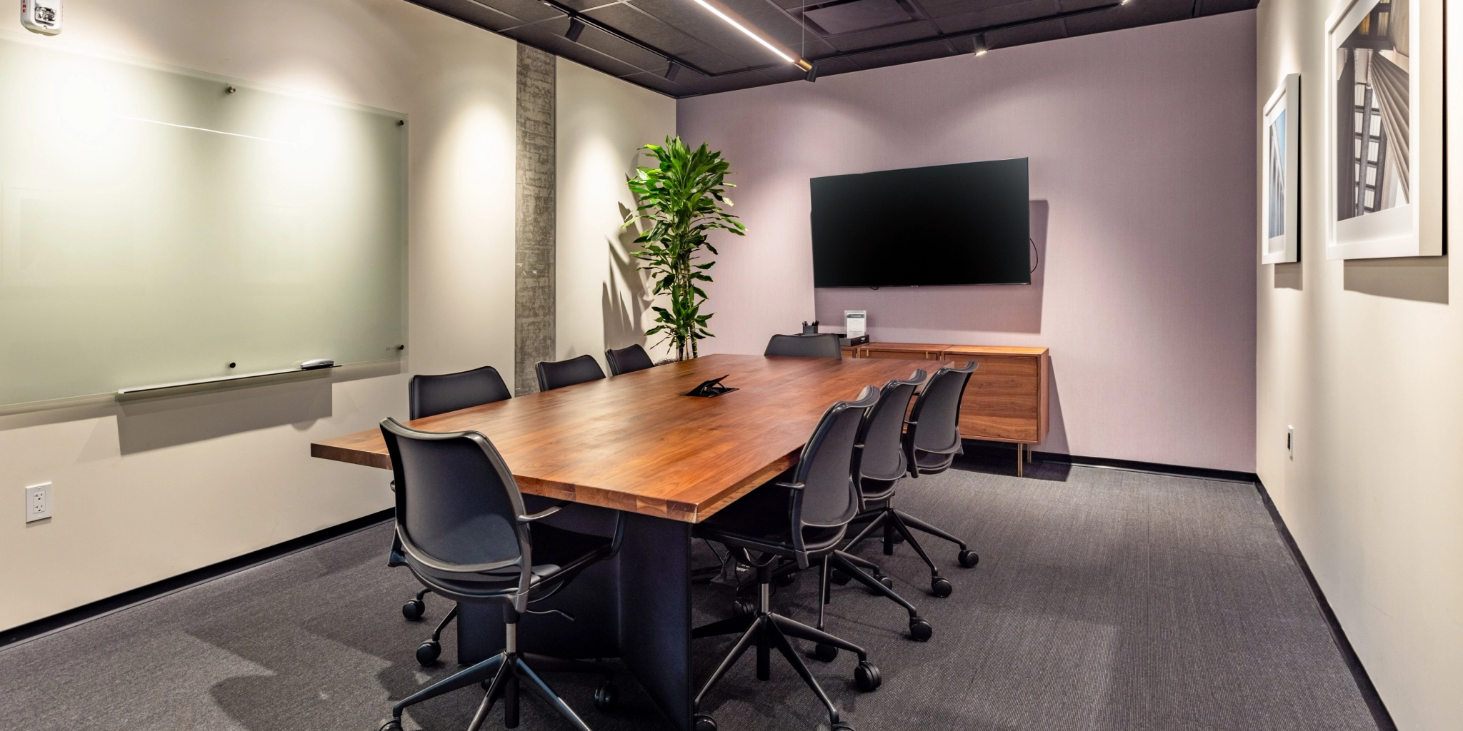 A modern coworking conference room in Washington features a wooden table, eight black office chairs, a large TV screen, a whiteboard, potted plant, and framed artwork on the walls.