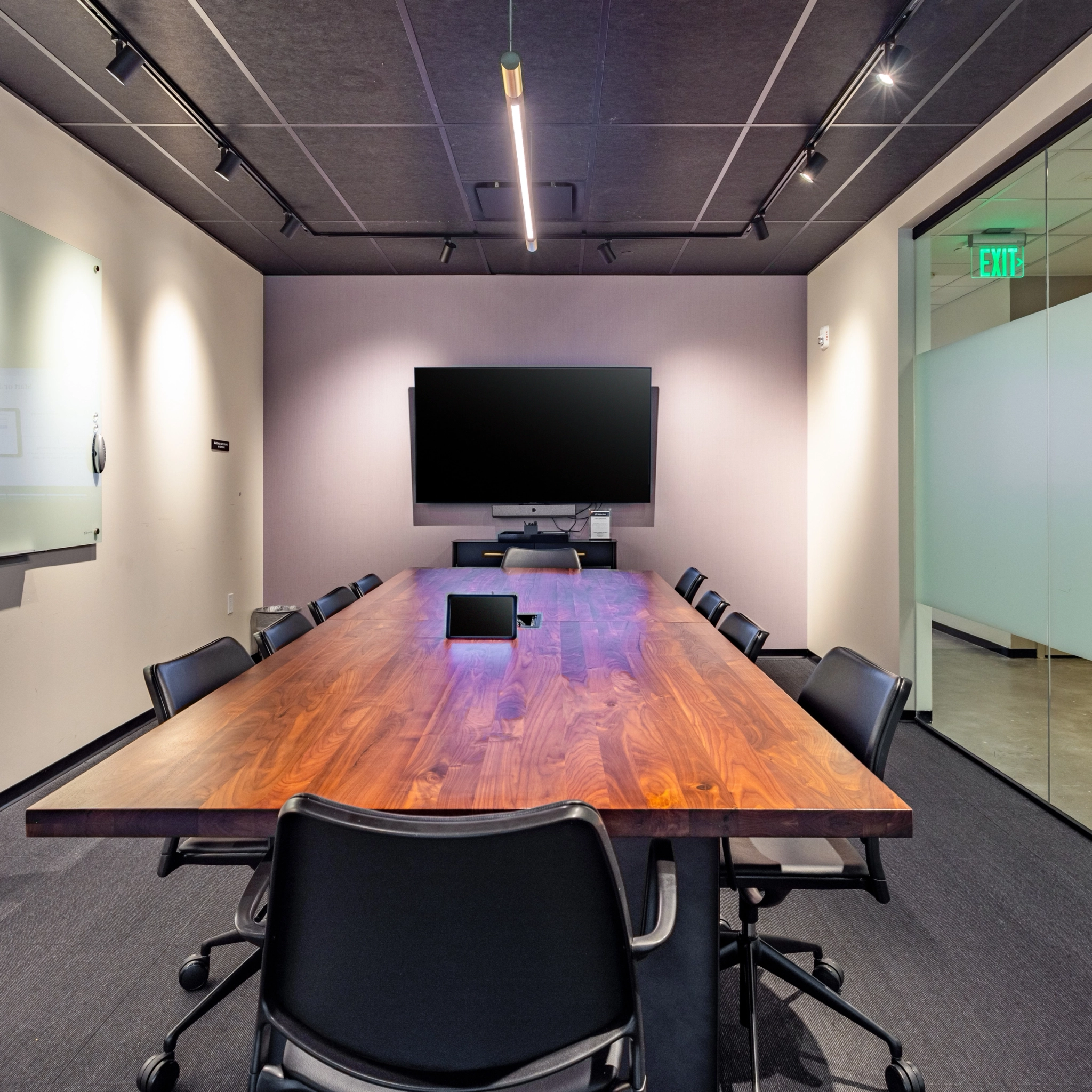 A modern meeting room with a large wooden table, black chairs, a wall-mounted TV, a glass panel door, and a wall-mounted whiteboard. Ideal for coworking in Washington.