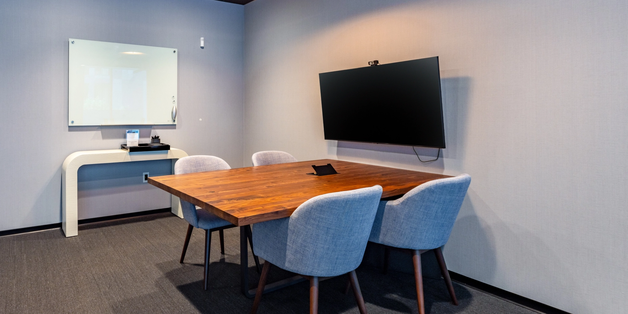 Een kleine coworking-vergaderruimte met een houten tafel, vier gevoerde stoelen, een aan de muur gemonteerde tv en een whiteboard.