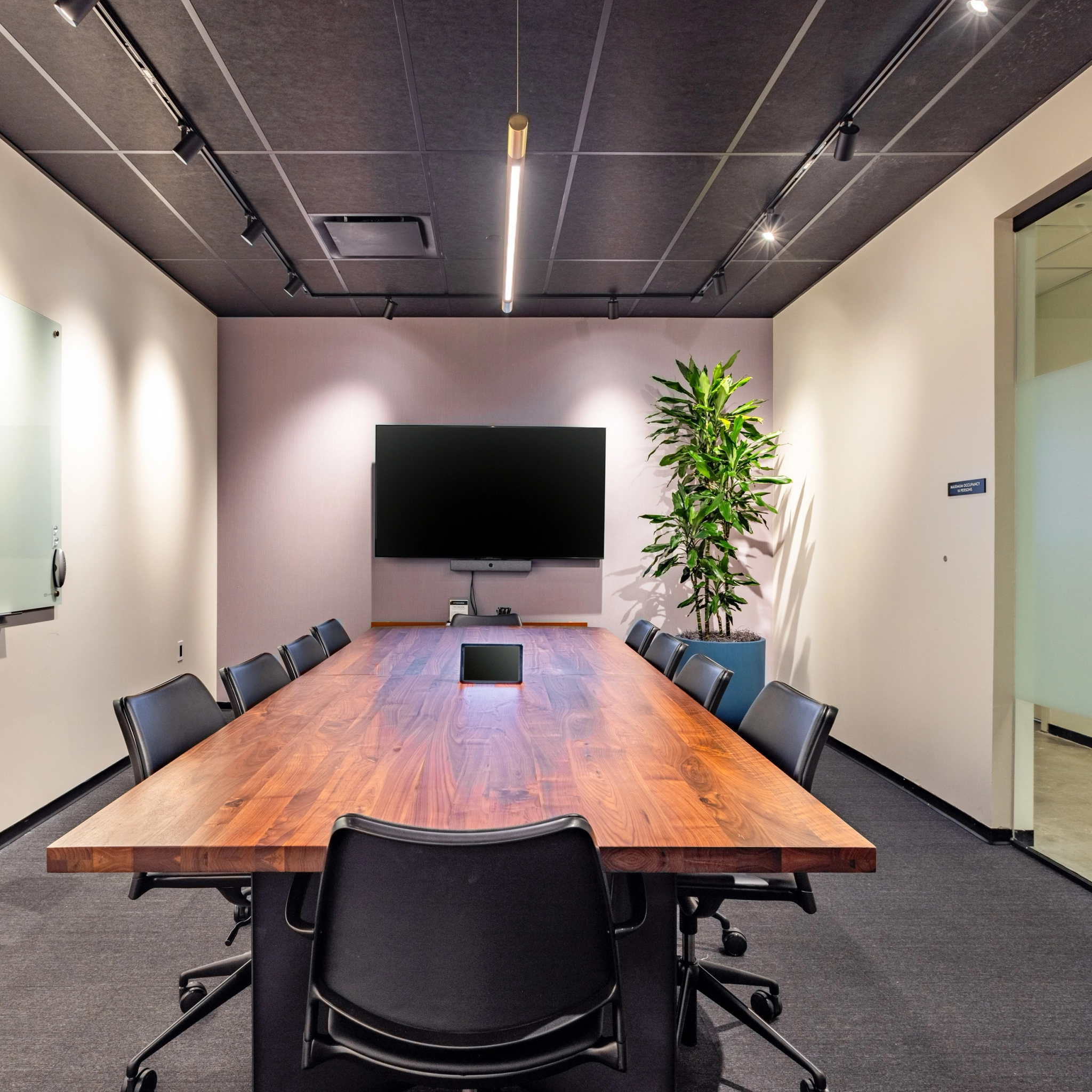 Une salle de conférence moderne dans notre bureau de Washington comprend une table en bois, neuf chaises noires, un grand écran mural, un tableau blanc en verre et une grande plante dans le coin, parfaite pour tout espace de travail productif.