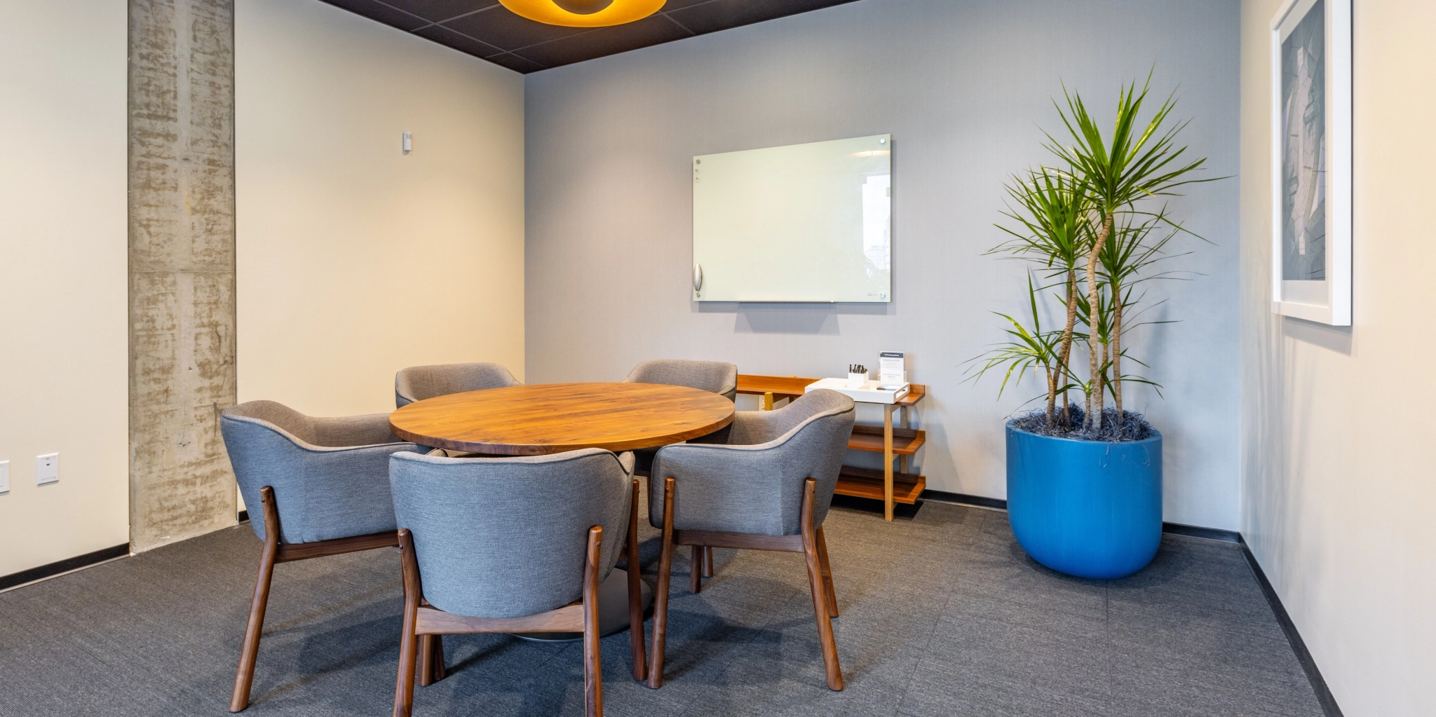 A small conference room in our Washington office, featuring a round wooden table, five gray chairs, a wall-mounted whiteboard, and a large potted plant in a blue planter, creates an ideal workspace.