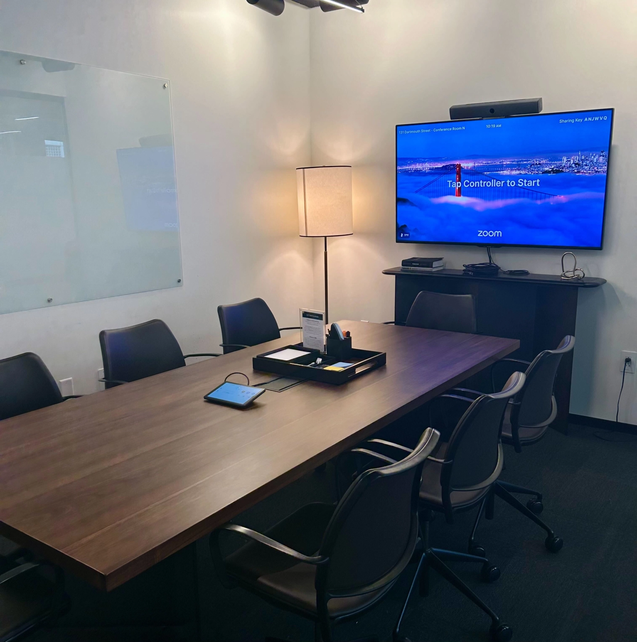 A modern office conference room with a large table, chairs, and a presentation screen displaying a zoom meeting interface.