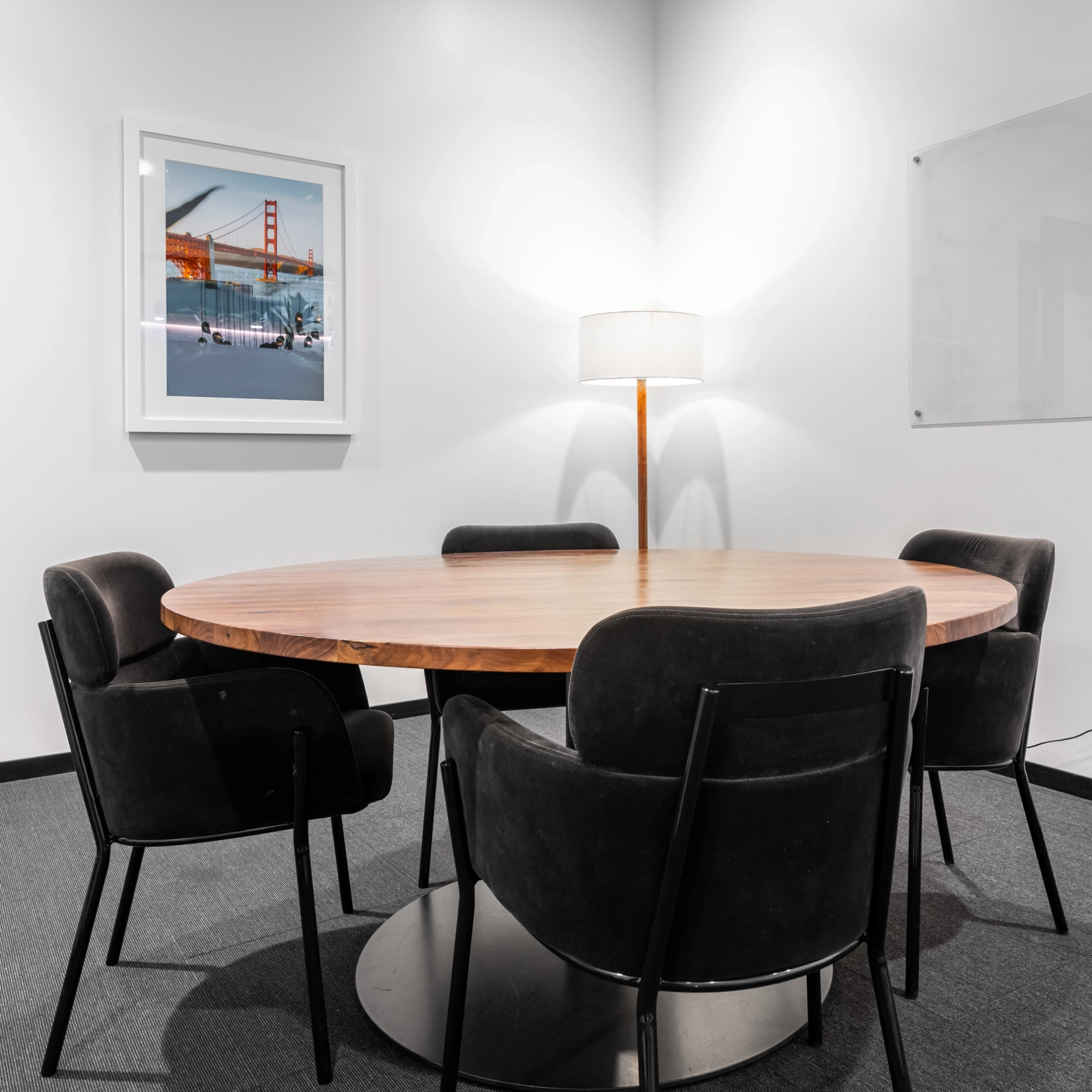 Een ronde houten tafel met vier zwarte stoelen creëert een gezellig hoekje in het Walnut Creek-kantoor. Een vloerlamp werpt zacht licht op een ingelijste foto van een brug, perfect voor inspirerende coworking-sessies.