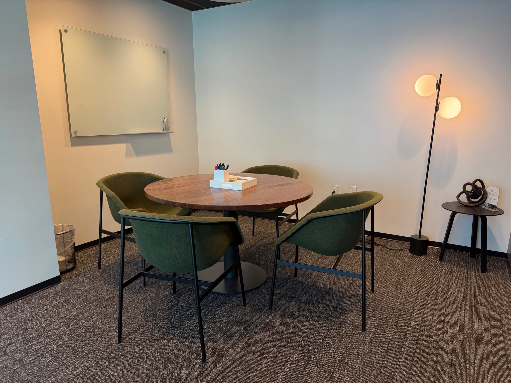 A cozy coworking meeting room featuring a round wooden table, four green chairs, a whiteboard, a floor lamp, and a side table with a decorative item—perfect for productive sessions in a modern workspace.