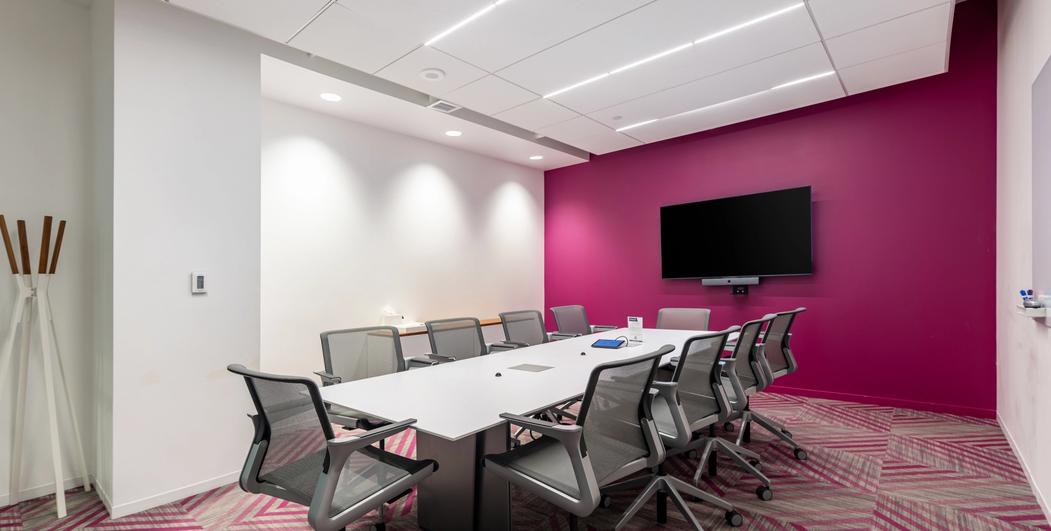 A modern coworking space in Arlington features a conference room with a large white table surrounded by gray office chairs. The back wall is painted magenta and has a mounted flat-screen TV. The workspace is well-lit and neatly arranged, perfect for meetings and collaboration.