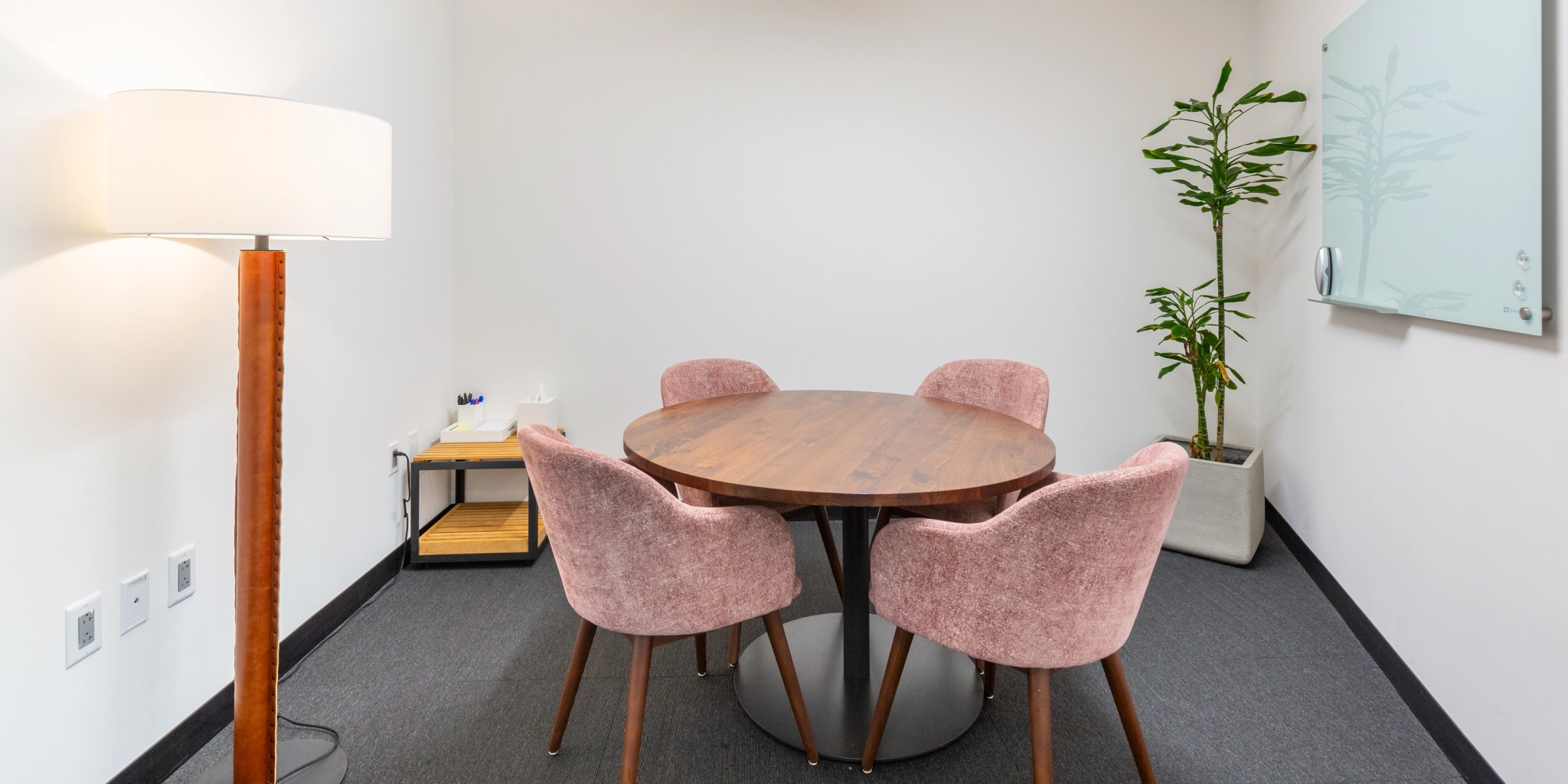 Small Arlington office meeting room featuring a round wooden table, four pink chairs, a floor lamp, a whiteboard, and a potted plant.