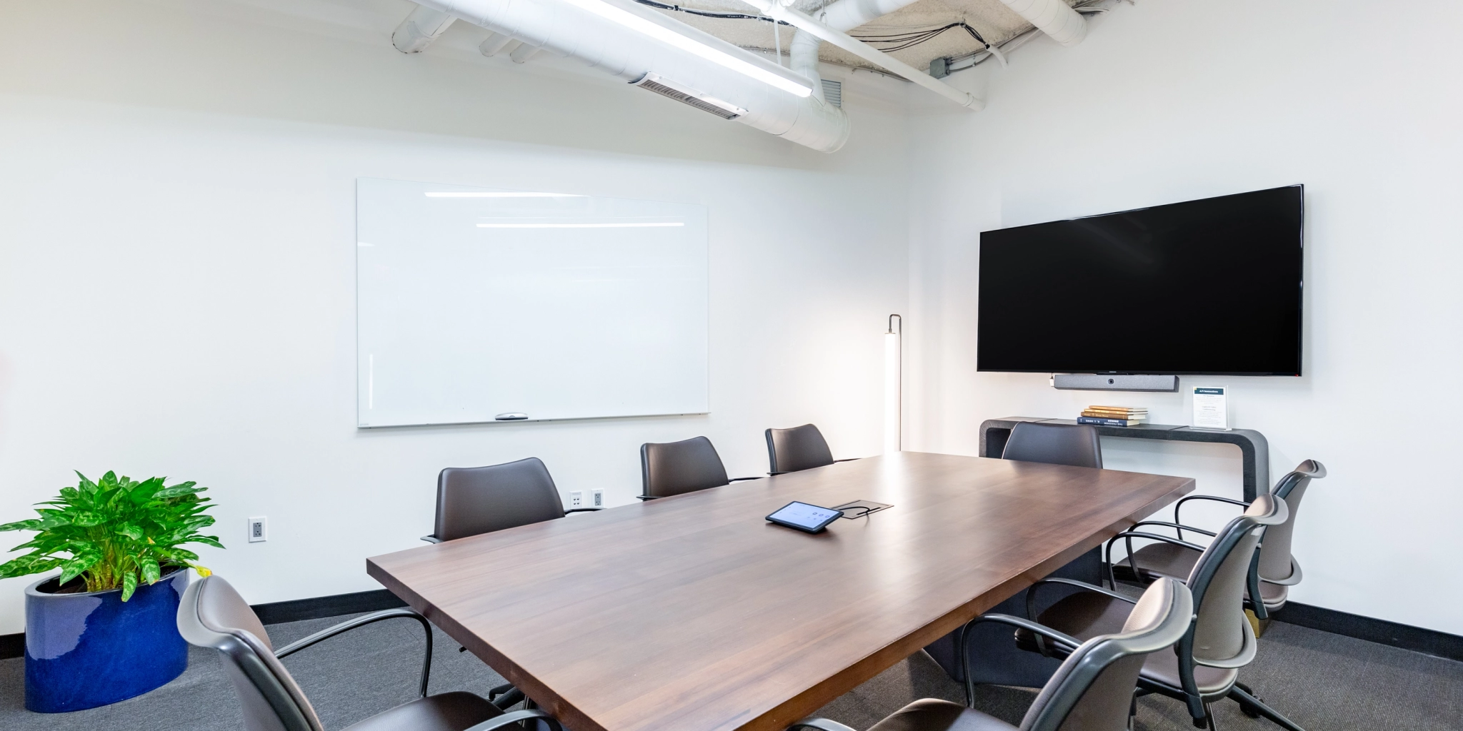 A coworking space featuring a wooden table with eight chairs, a wall-mounted TV, and a whiteboard. The meeting room is completed with a plant in the corner and a tablet resting on the table.