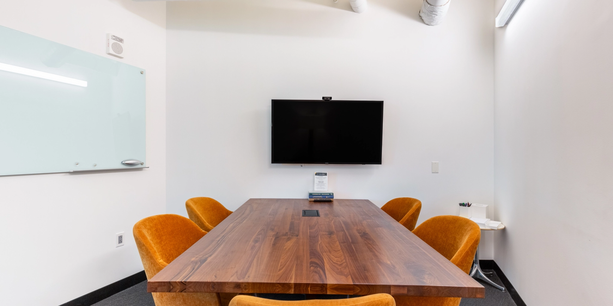 A cozy meeting room in the Arlington office features a wooden table, six orange chairs, a wall-mounted TV, and a whiteboard. Perfect for collaborative workspaces!