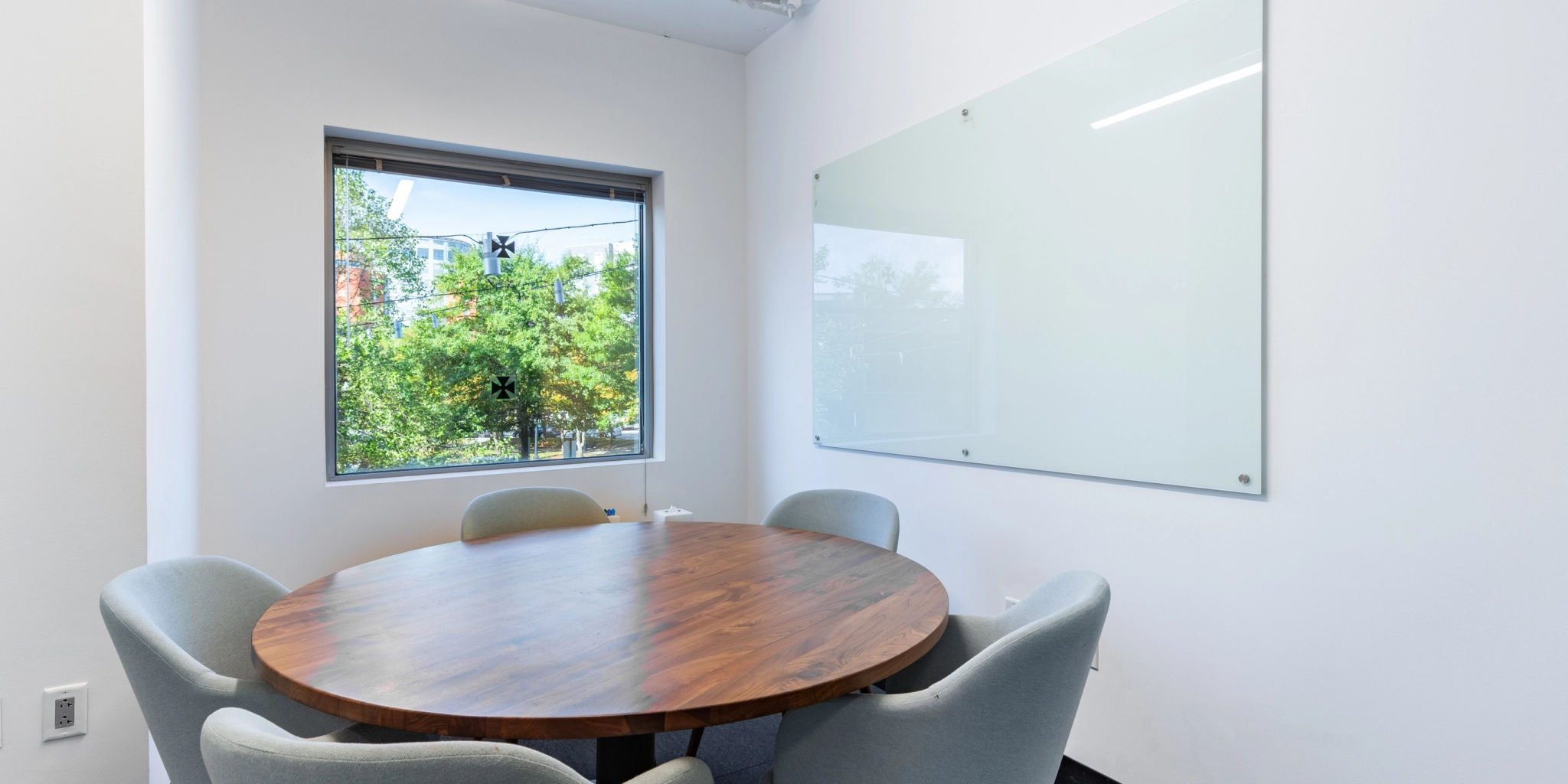 A cozy Arlington workspace, this small conference room features a round wooden table, four gray chairs, and a large blank whiteboard on the wall. The window offers a refreshing view of greenery outside, perfect for fostering creativity and collaboration.