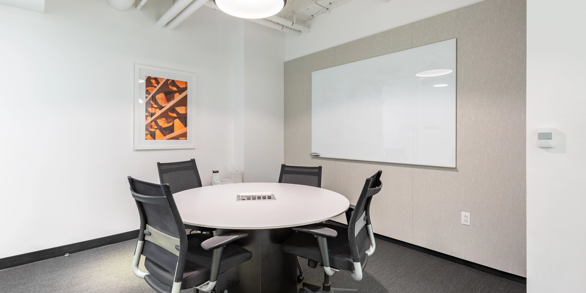 A small meeting room in the Arlington office features a round table, four chairs, a whiteboard on the wall, and a framed abstract art piece.