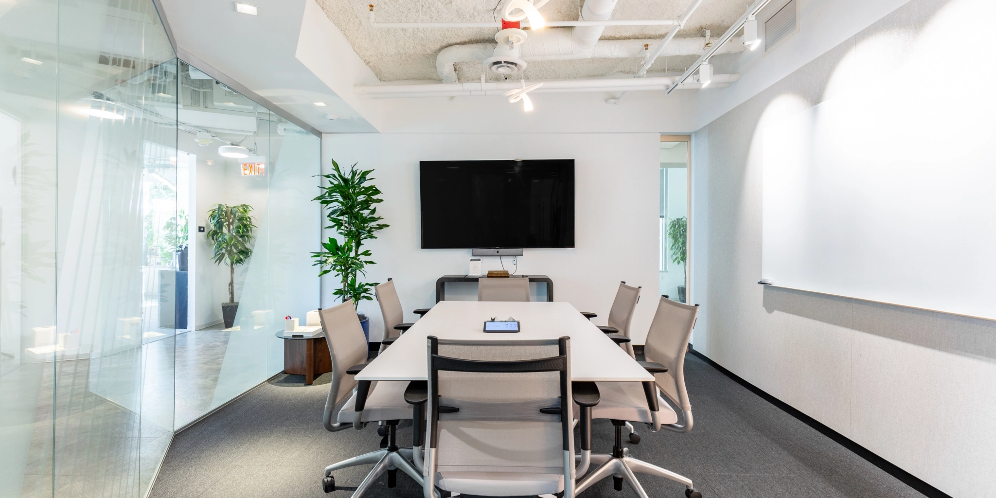 Un espace de coworking moderne avec une longue table, huit chaises et un grand écran mural. Les murs en verre créent une sensation d'ouverture, tandis que les plantes décoratives rehaussent l'ambiance de l'environnement de bureau.