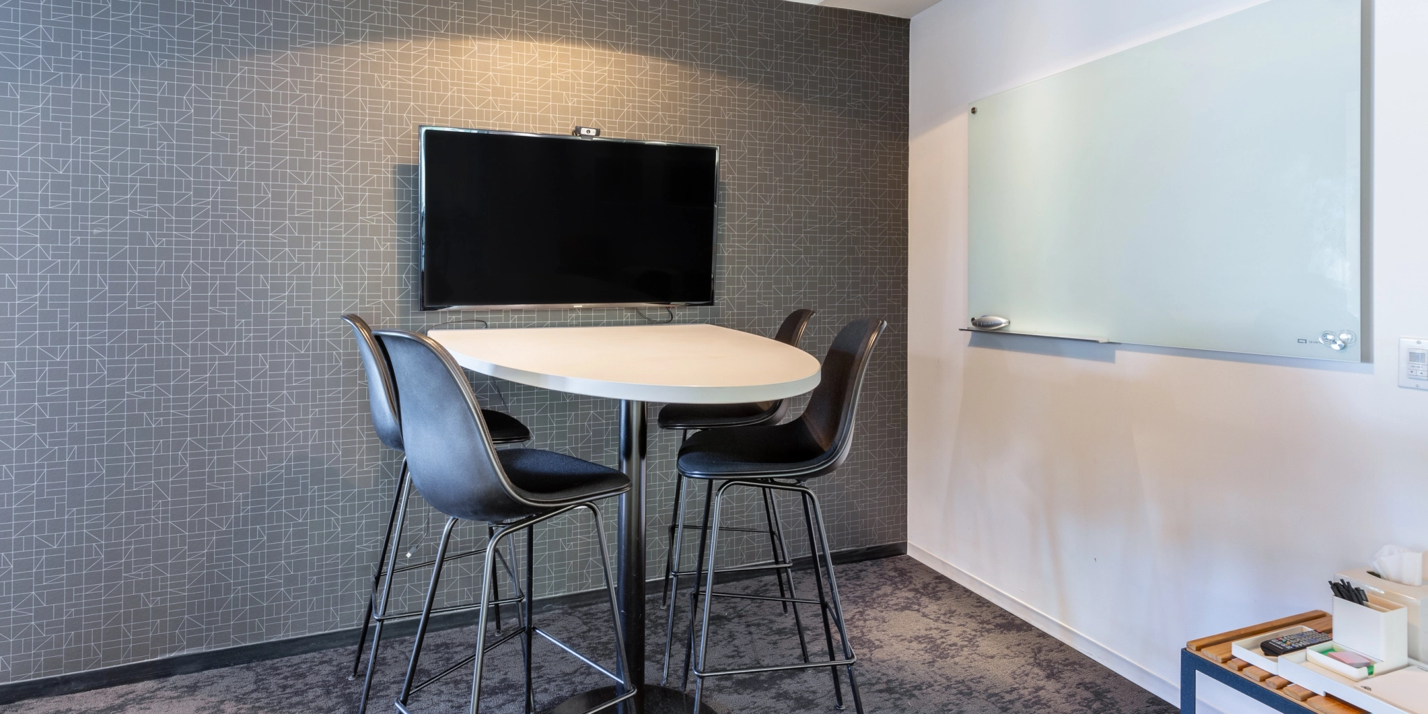 A cozy coworking space in Arlington, featuring a meeting room with a round table, four high chairs, a wall-mounted TV, and a whiteboard.