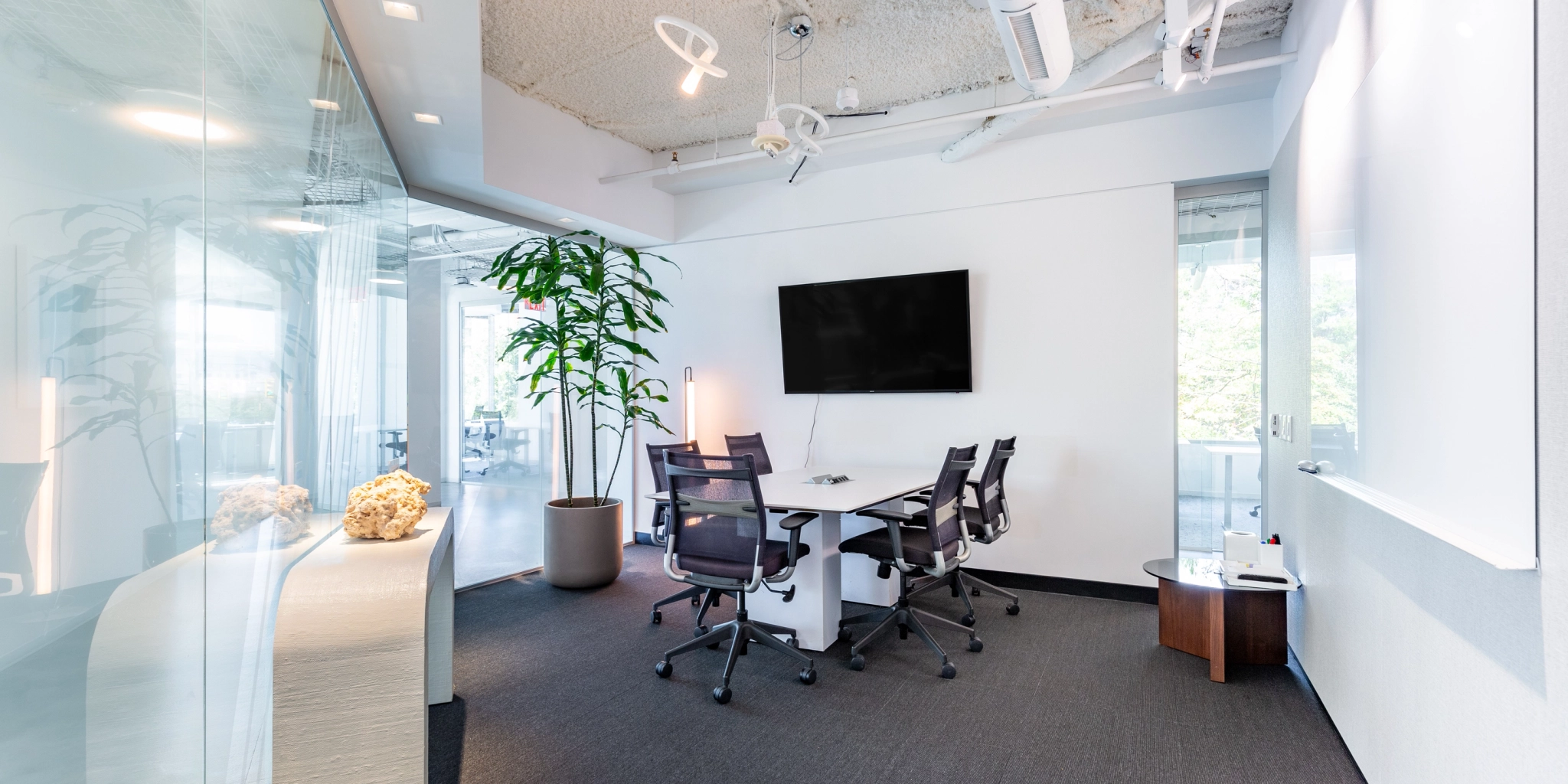 A modern coworking space with a small meeting area, featuring three chairs around a table, a wall-mounted screen, a large plant, and glass walls.