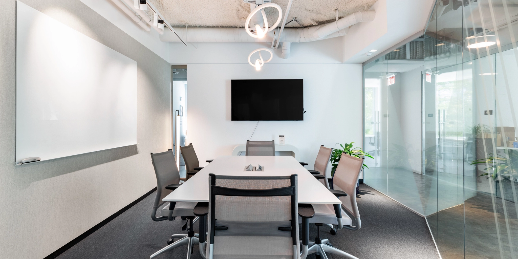 Een moderne vergaderruimte in een kantoor in Arlington is voorzien van een lange tafel, acht stoelen, een aan de muur gemonteerde tv en een whiteboard dat wordt verlicht door overheadverlichting. Glazen deuren en een weelderige plant versterken de elegante sfeer.