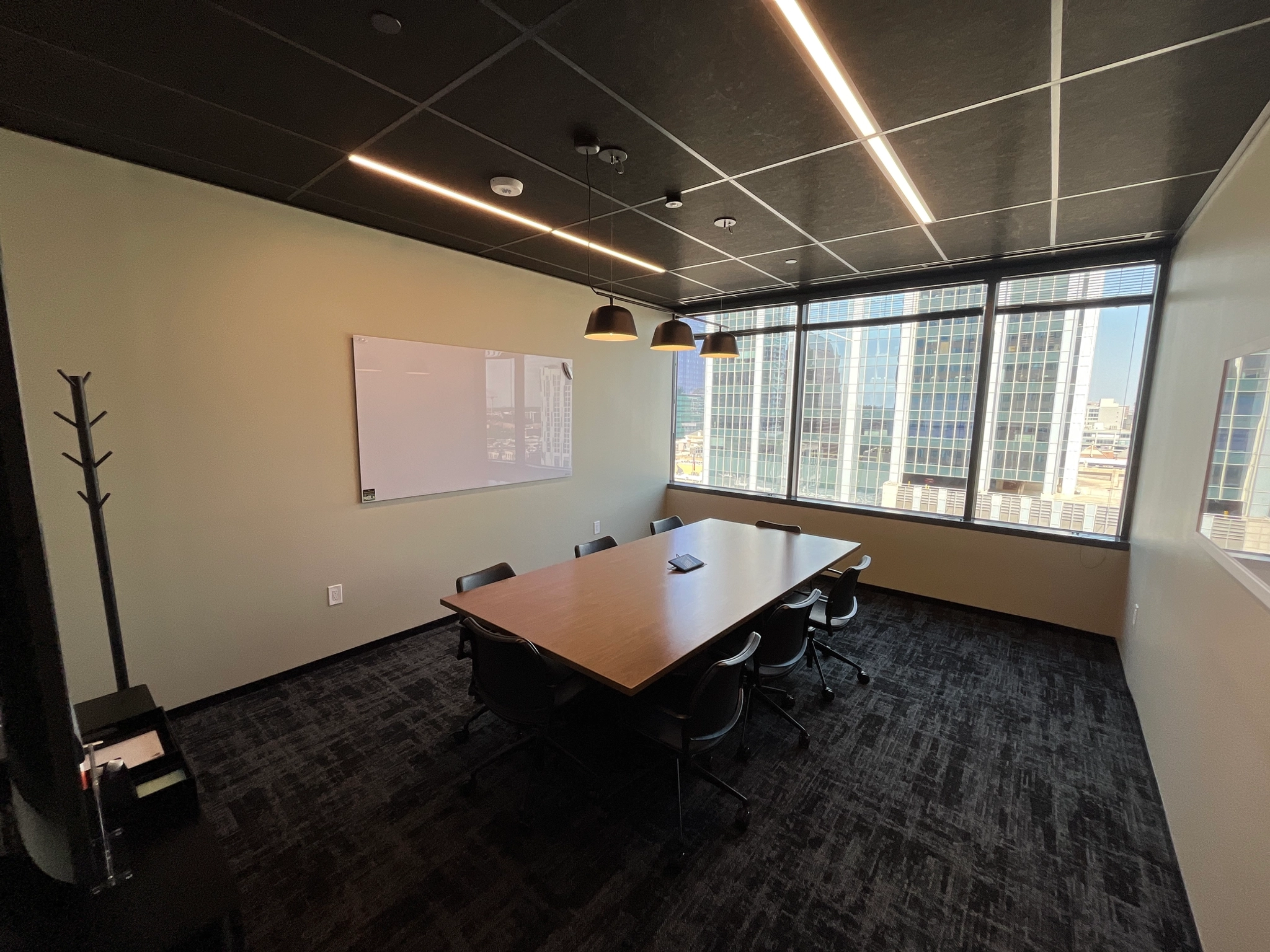 A modern meeting room with a long table, six chairs, a whiteboard on the wall, ceiling lights, and large windows offering a view of skyscrapers.