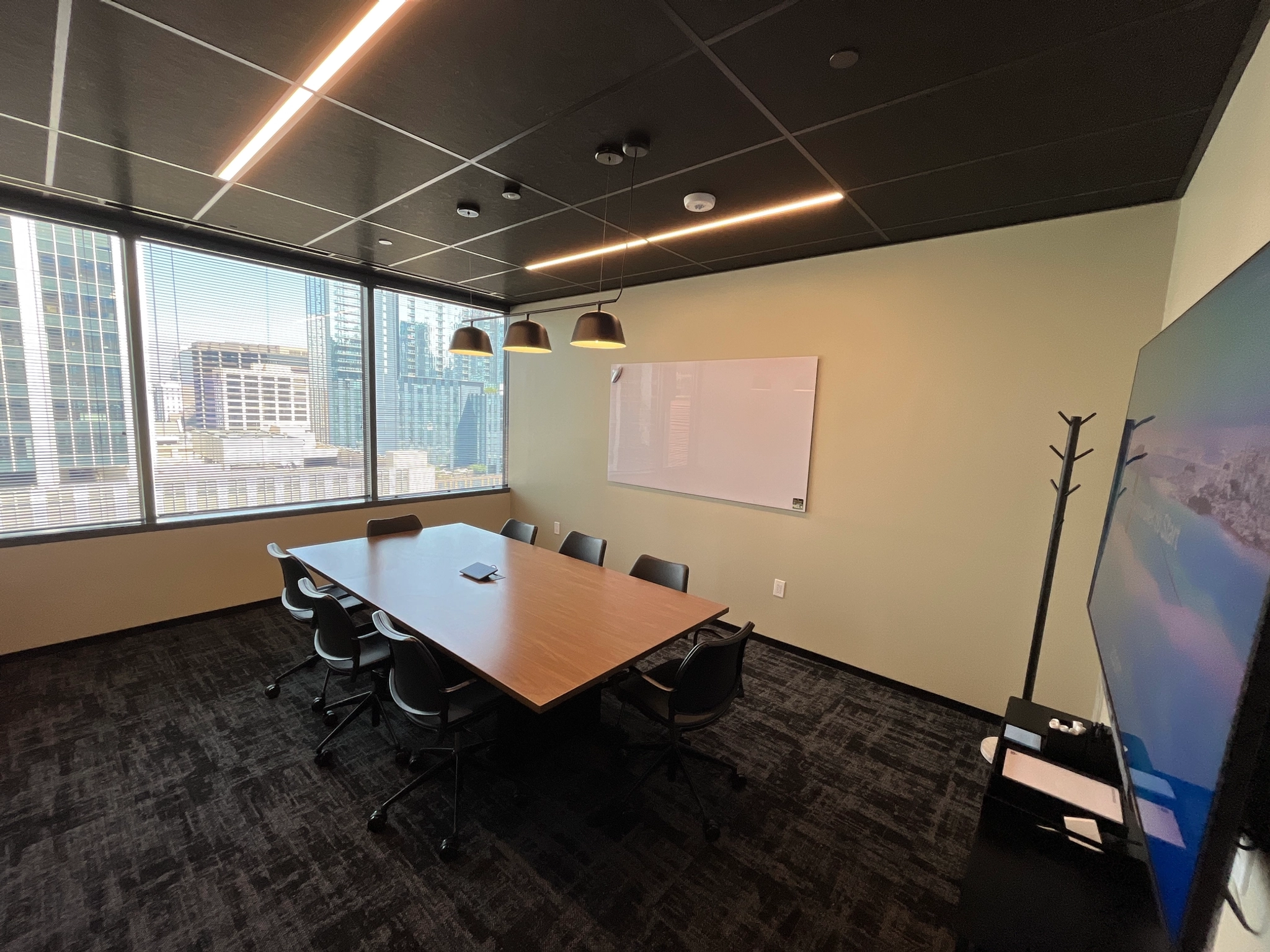 A modern meeting room featuring a long table, eight chairs, large windows showcasing a cityscape view, a whiteboard, pendant lights, and a TV on the right wall. This versatile workspace is ideal for collaboration and creativity.