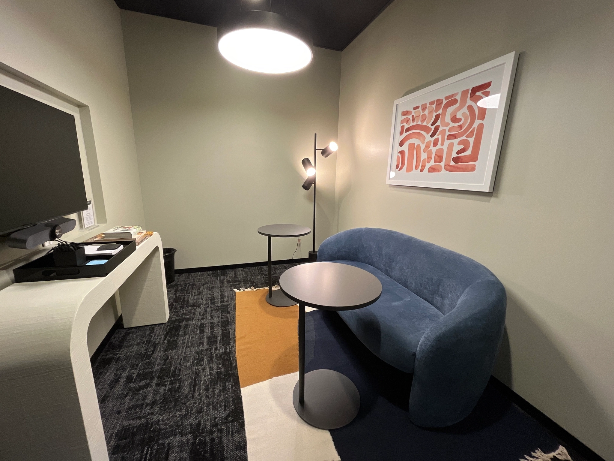 Een moderne coworking-lounge in Austin beschikt over een blauwe bank, twee ronde tafels, een staande lamp, een tv en abstracte kunst aan de muur.