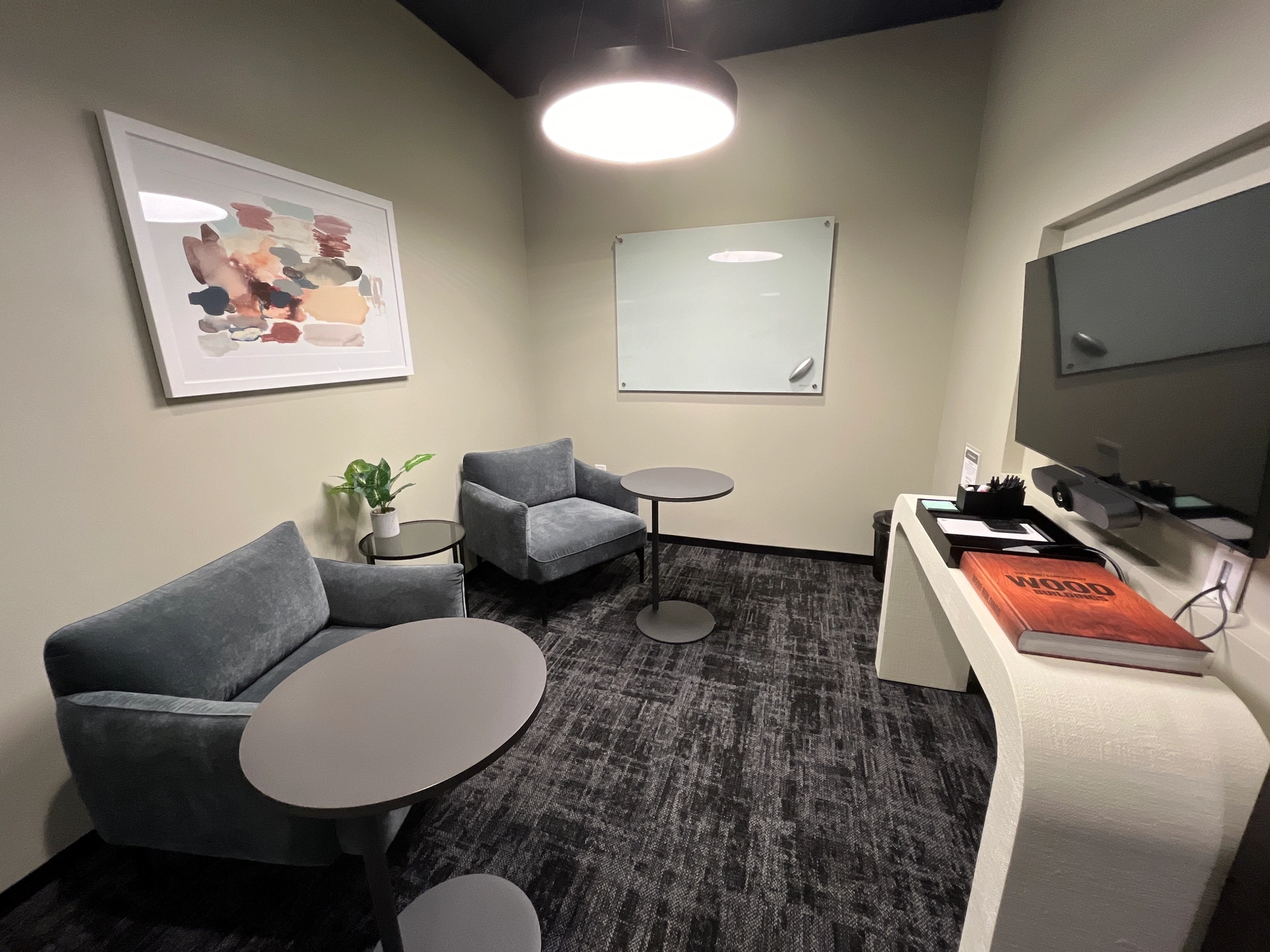 A cozy meeting room featuring two gray chairs, two round tables, a wall-mounted TV, a whiteboard, and an abstract art piece. This workspace also includes a plant in the corner for a touch of greenery.