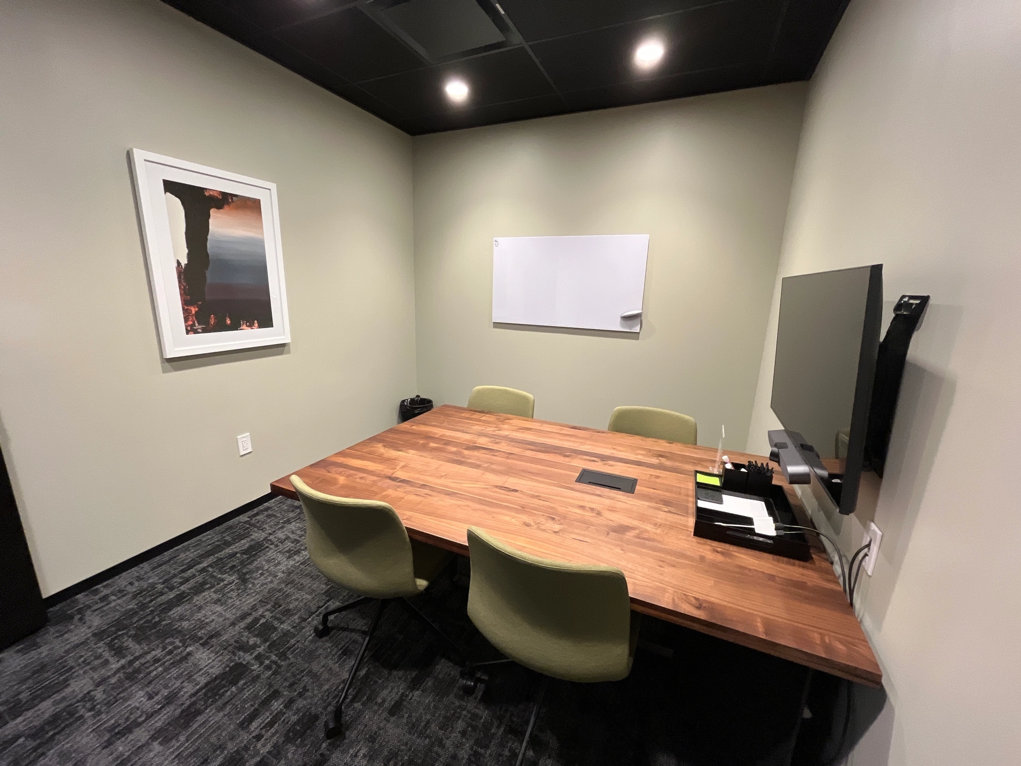 Een gezellige coworking-vergaderruimte in Austin, met een houten tafel, drie groene stoelen, een aan de muur gemonteerde tv, een whiteboard en een ingelijste foto.