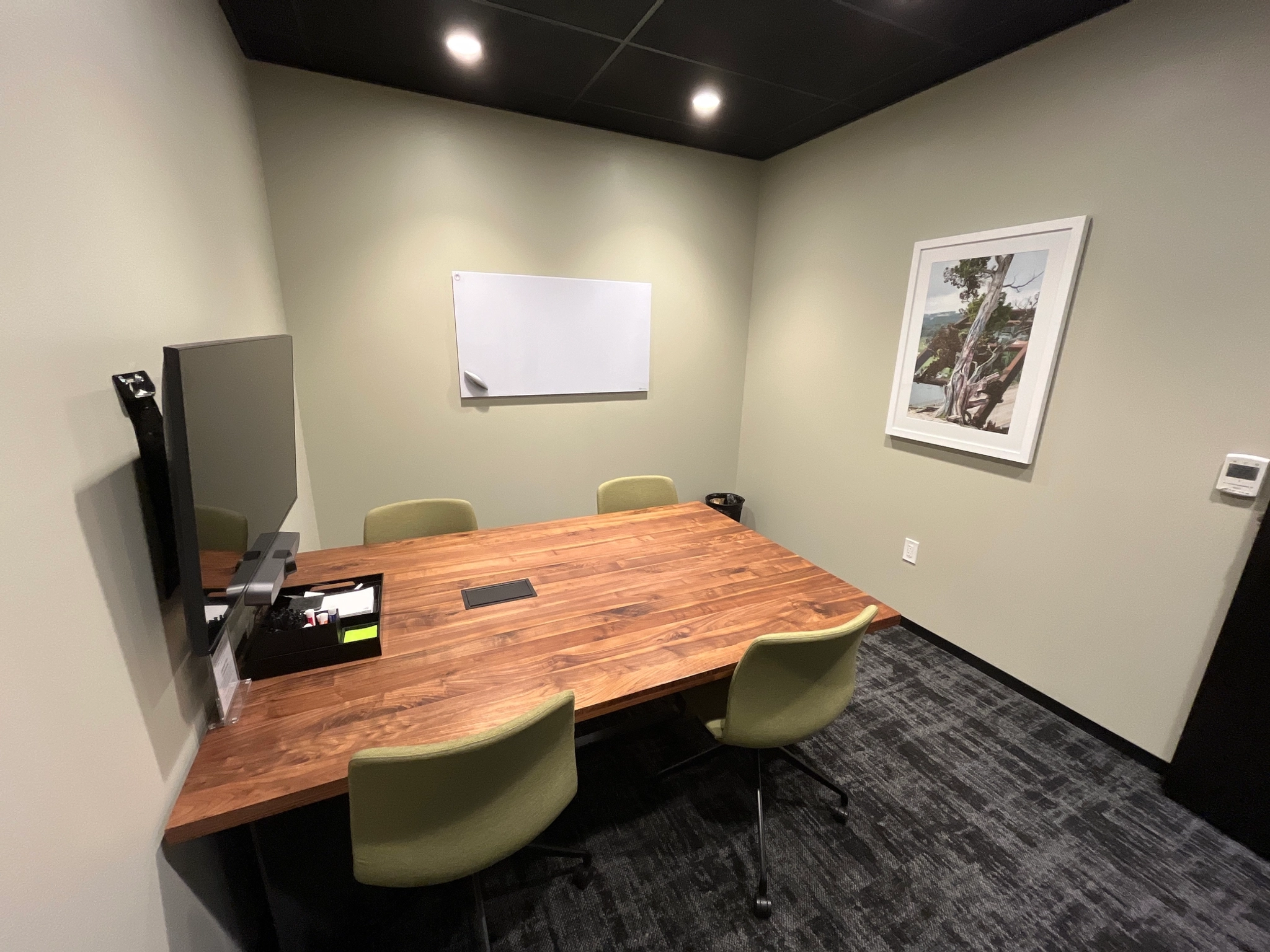 Een kleine coworking-conferentieruimte in Austin beschikt over een houten tafel, vier groene stoelen, een aan de muur gemonteerde tv, een leeg whiteboard en ingelijste kunst aan de muur.