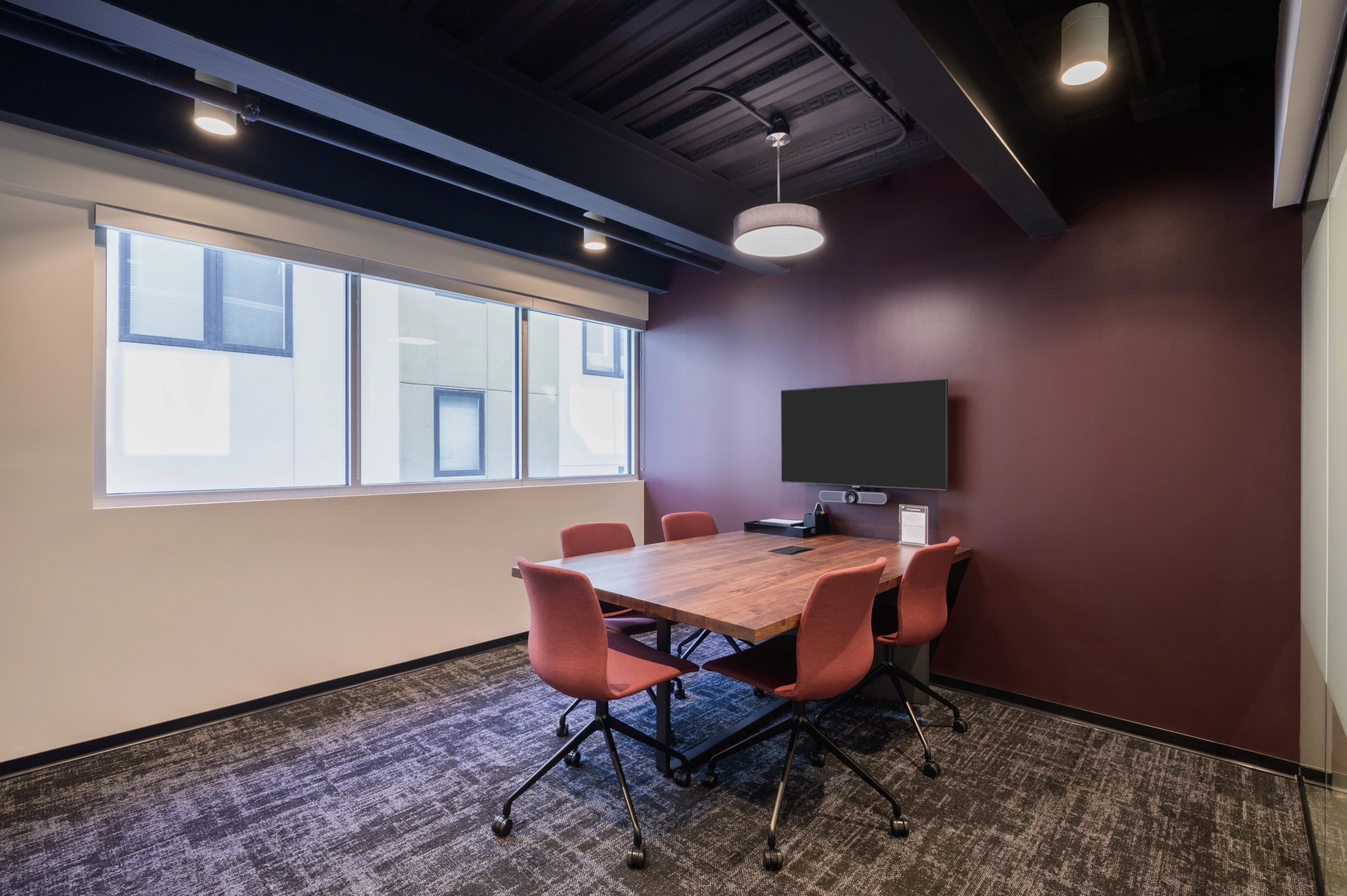 Modern coworking meeting room with a wooden table, red chairs, a television display, and natural light from a window in Denver.