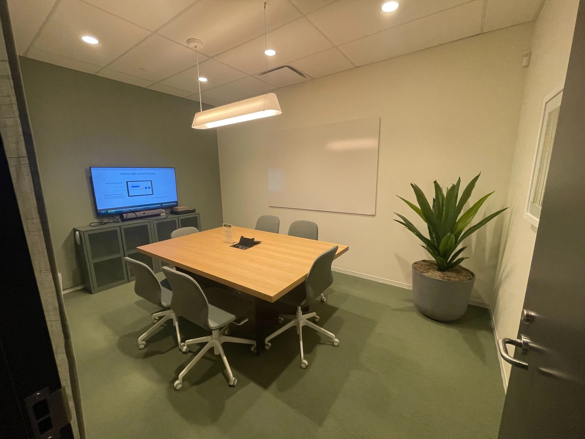 Une salle de conférence de bureau moderne équipée d'une table en bois, de quatre chaises, d'un grand moniteur, d'un tableau blanc et d'une plante en pot dans le coin.