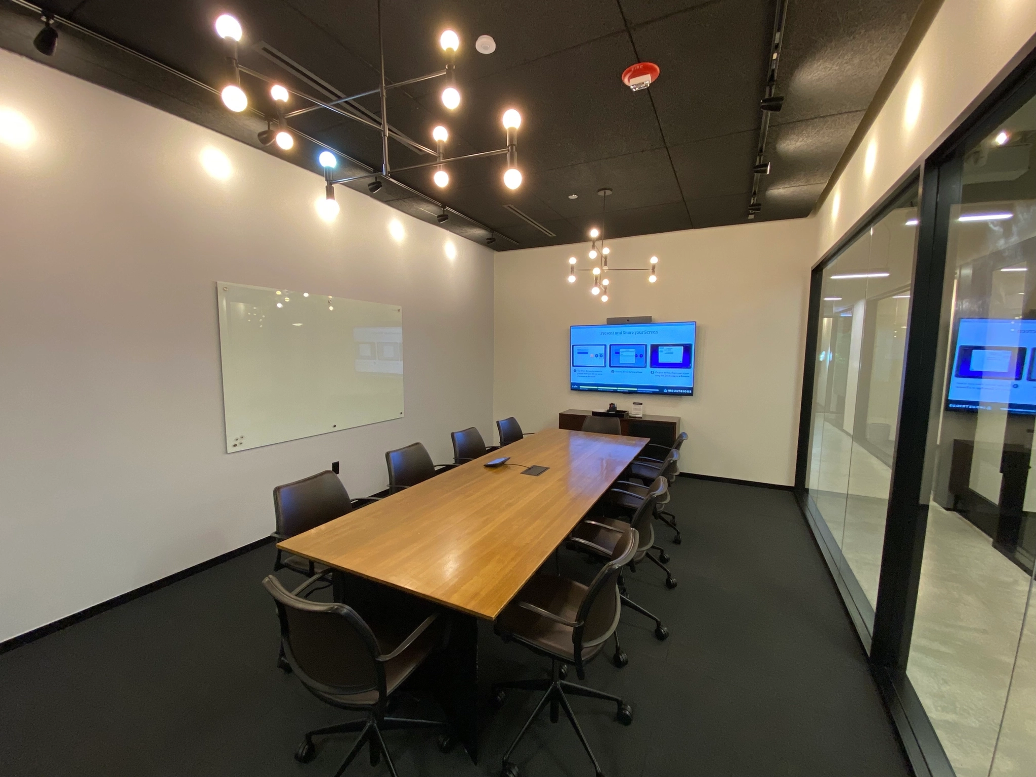 A coworking space featuring a conference room with a long wooden table, eight chairs, and modern lighting fixtures. A whiteboard complements the wall-mounted screen displaying presentations, creating an ideal workspace for collaboration.