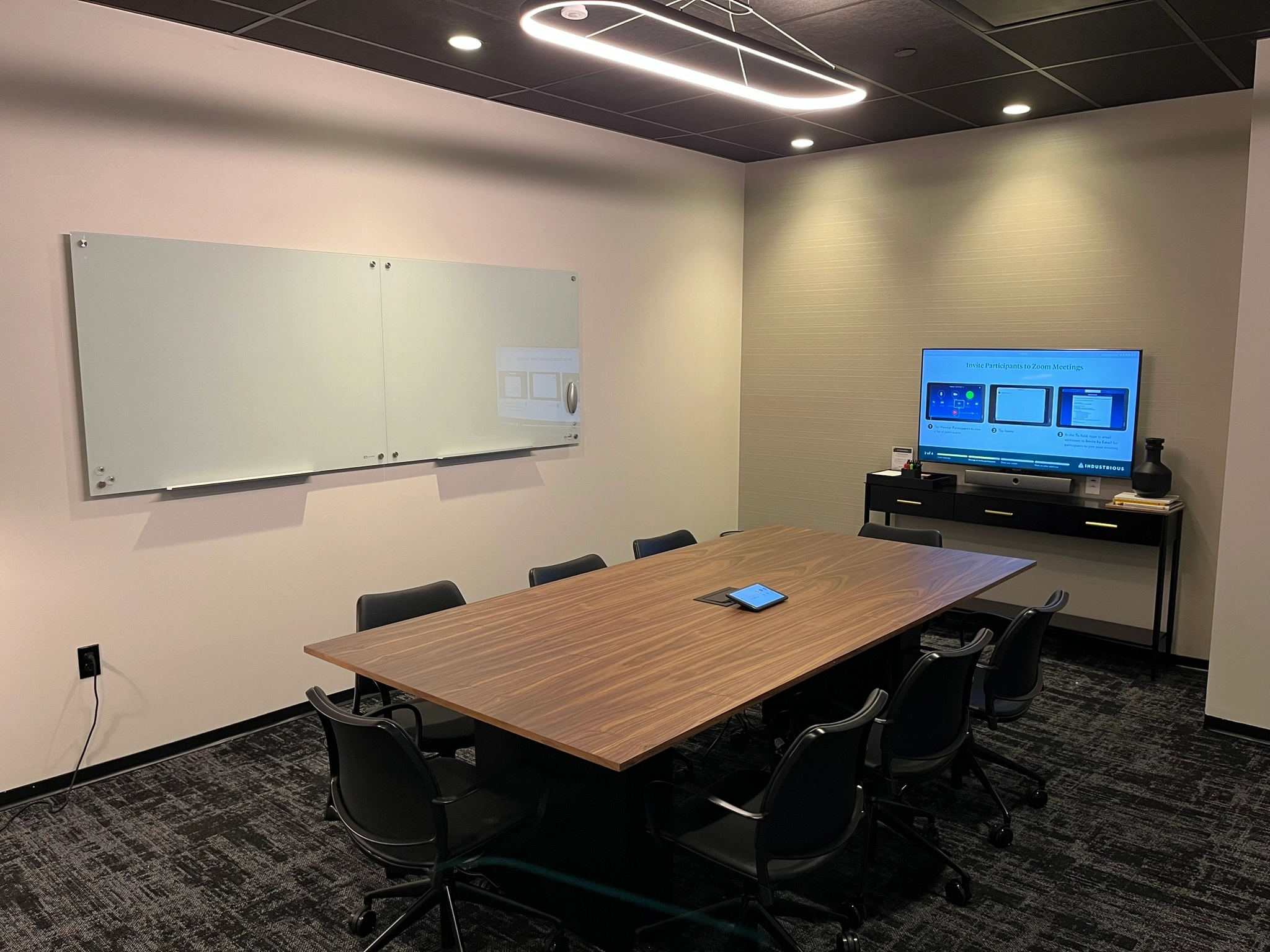 Modern meeting room with a large table, chairs, dual monitor setup, and whiteboards.