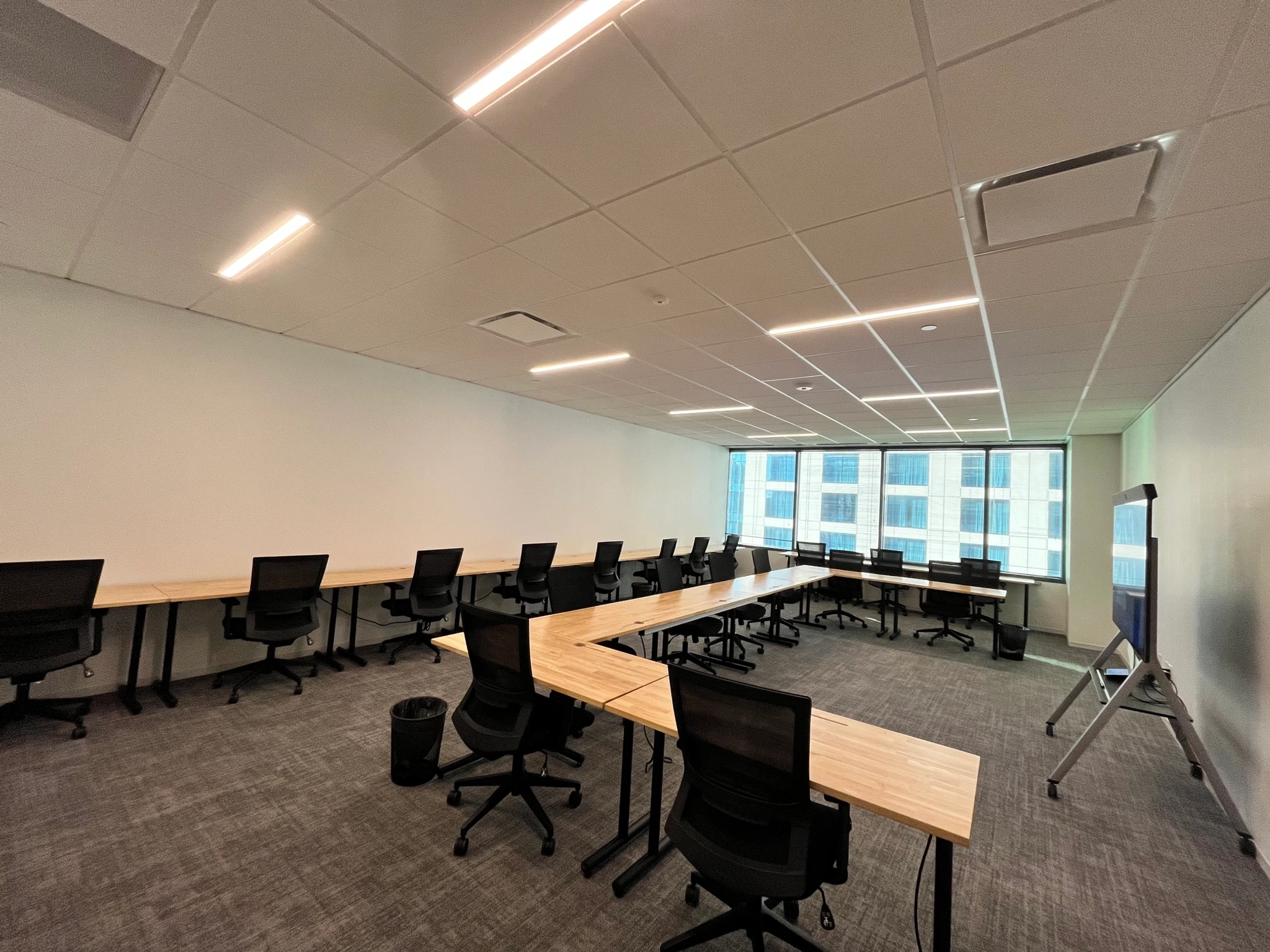 A modern office space in Austin with a U-shaped arrangement of wooden desks and black office chairs. There is a large window on one side, and a whiteboard on a stand is on the right side of the meeting room.