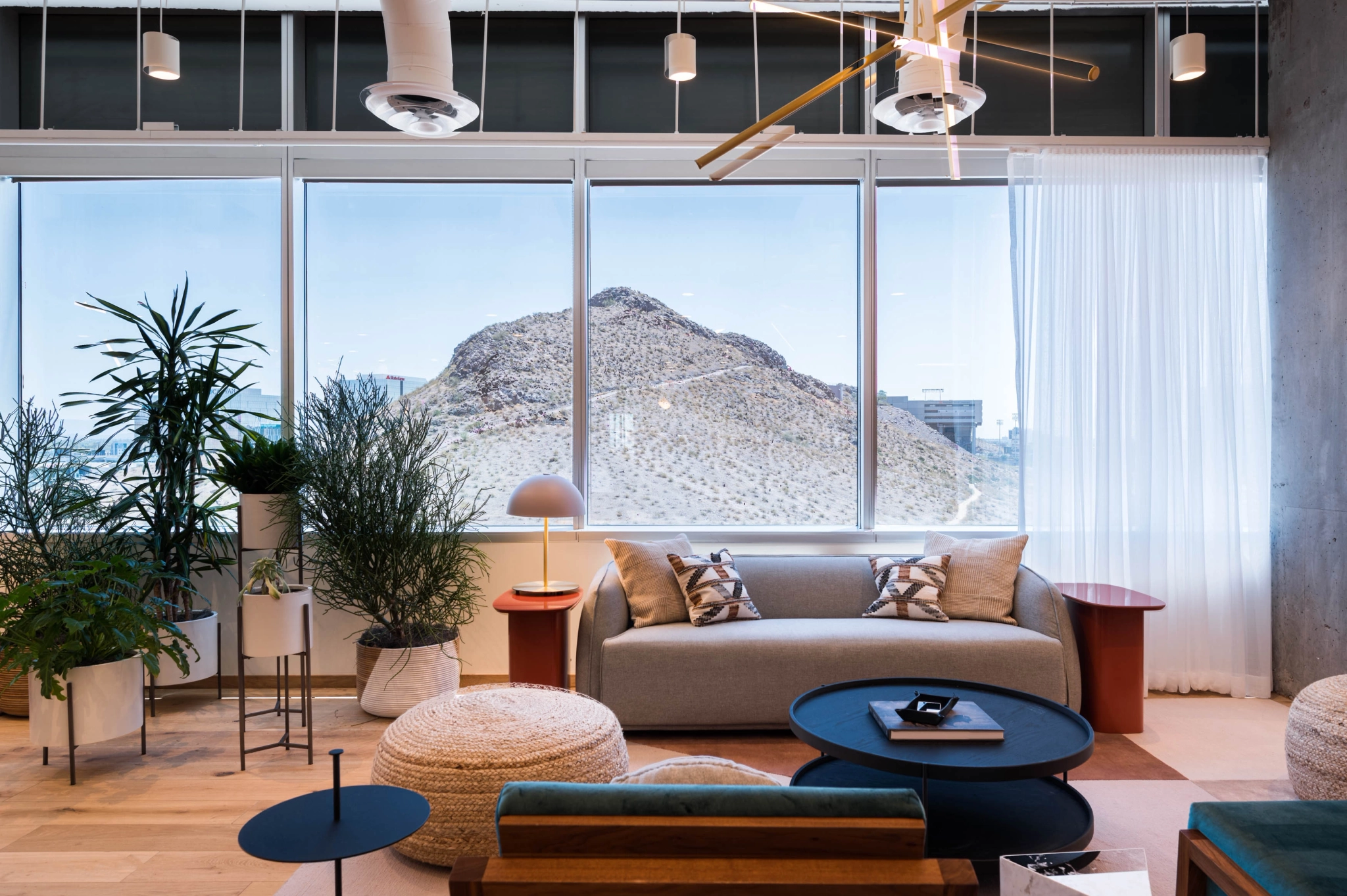 Modern coworking lounge with a sofa, armchairs, and tables, overlooking a mountain through large windows, flanked by indoor plants.