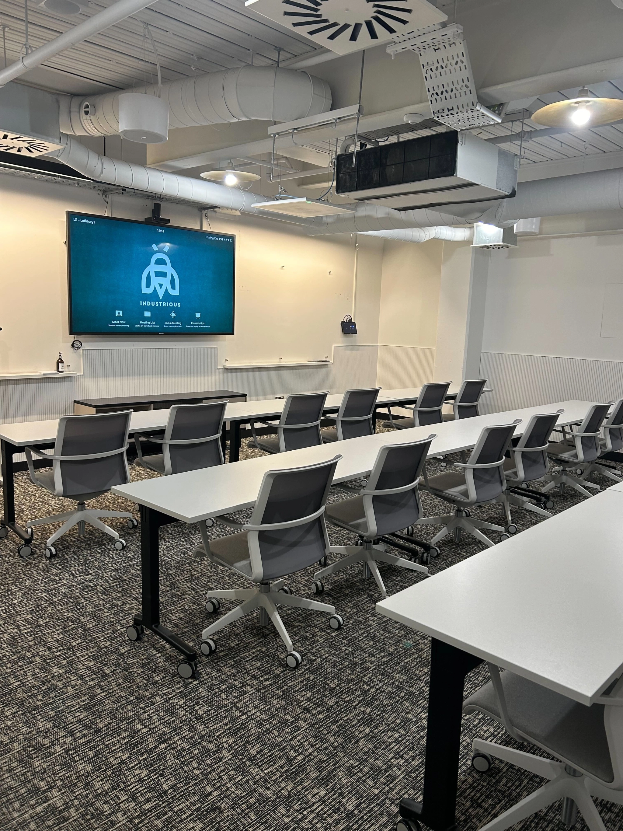 A modern coworking space in London features a conference room with rows of white tables and gray chairs, all facing a large wall-mounted screen displaying a presentation.