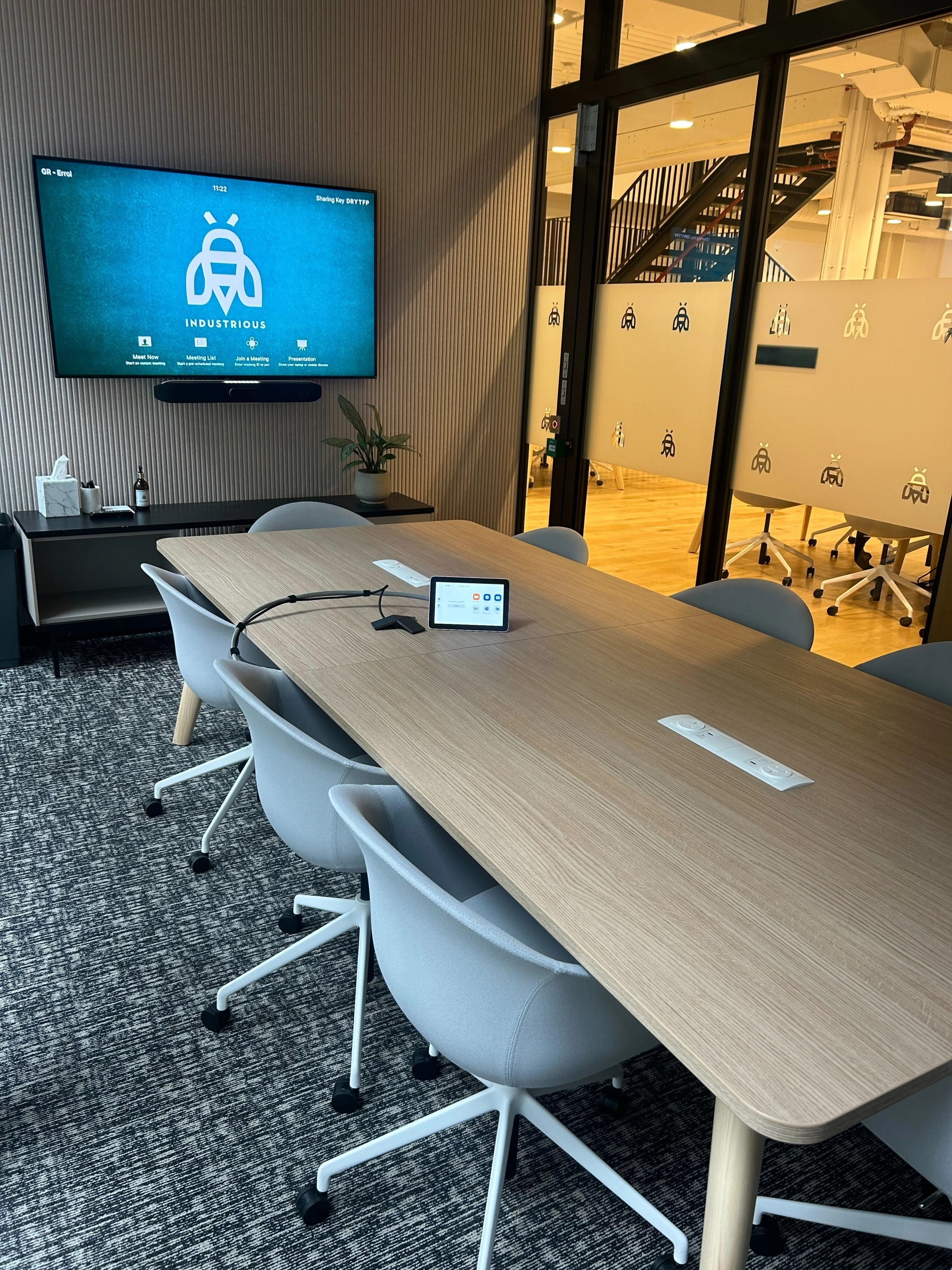 A modern meeting room with a large table, several gray chairs, and a wall-mounted screen displaying a logo, all within a coworking space in London divided by a glass wall from another workspace.