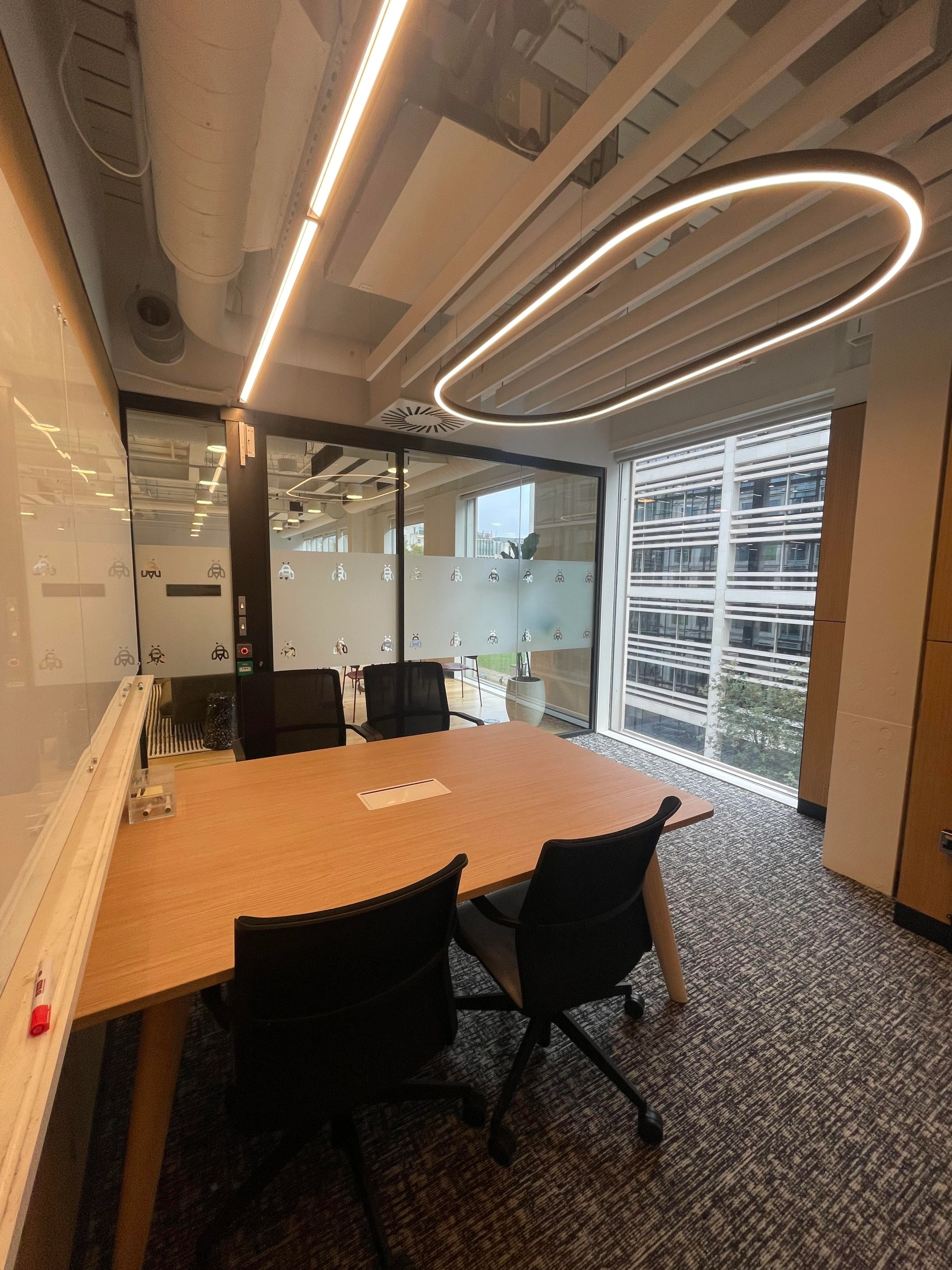 Modern meeting room featuring a wooden table, black chairs, and a large ring-shaped ceiling light. A glass wall offers a view of a building exterior. Whiteboard and marker are on the left, creating an efficient workspace for collaboration.