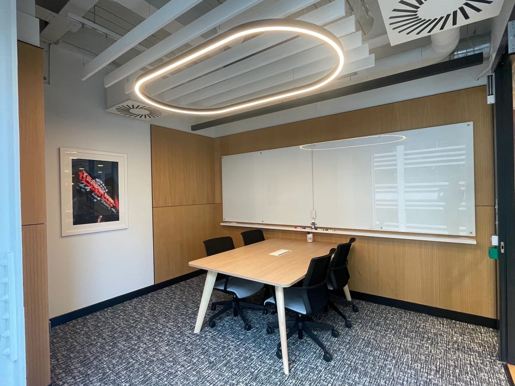 A modern workspace in London featuring a wooden table, five chairs, large whiteboard, abstract art adorning the wall, and an oval ceiling light fixture. Perfect for coworking and collaborative efforts.