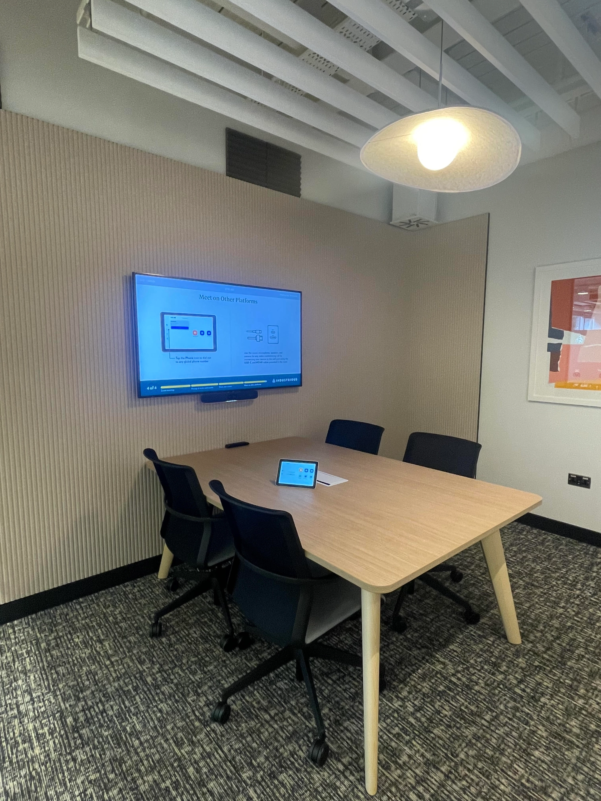 Salle de conférence de bureau avec une table en bois, quatre chaises noires, un écran mural diffusant une présentation et un plafonnier à Londres. Une tablette repose sur la table, la rendant prête à répondre à tous les besoins de la salle de réunion.