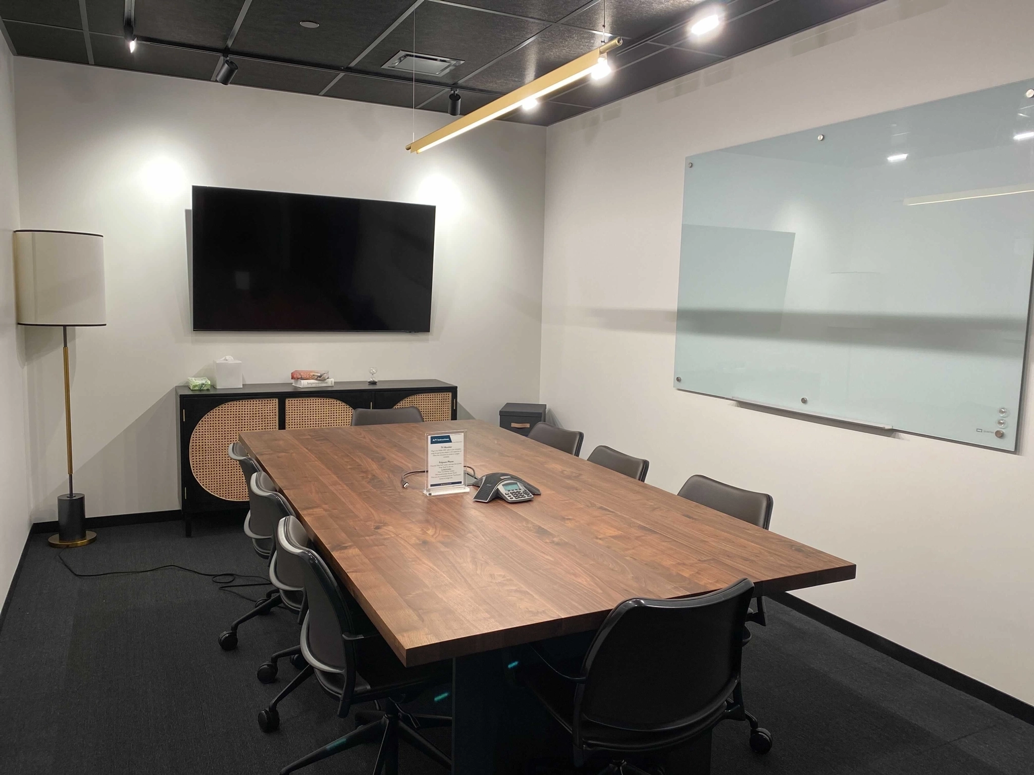 Modern coworking conference room in Washington featuring a wooden table, eight chairs, a wall-mounted TV, whiteboard, floor lamp, and side cabinet. A phone and a sign rest on the table—perfect for your next meeting room needs.