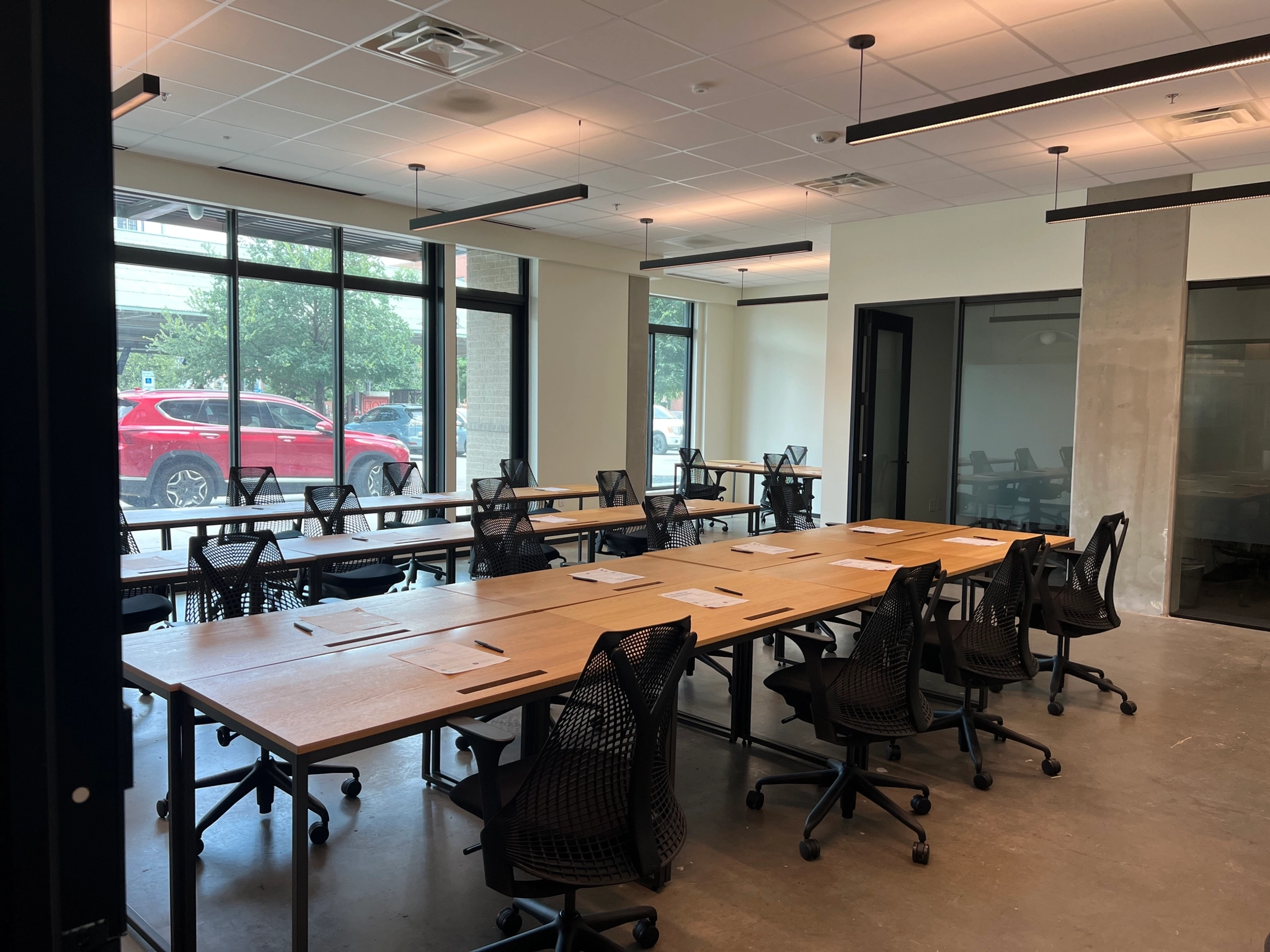 Un espace de coworking moderne avec des rangées de bureaux et de chaises. De grandes fenêtres offrent de la lumière naturelle et une vue sur une voiture rouge garée à l'extérieur dans le quartier animé de Dallas.